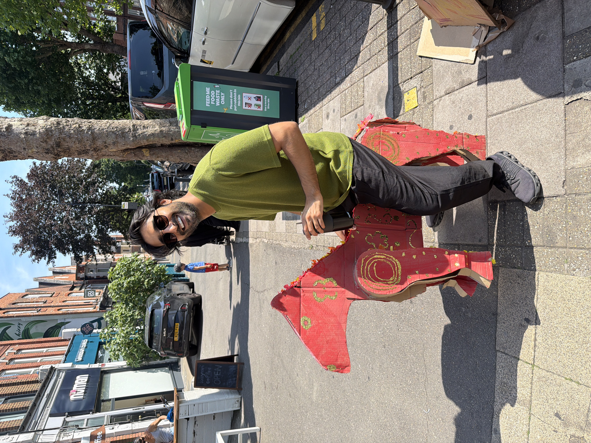 A man with dark hair and sunglasses is standing on a sidewalk next to a red cardboard cutout of a horse. He is wearing a green t-shirt and black pants, and he is holding a black travel mug. The horse cutout is decorated with gold glitter and has a fringe of red and orange material around its head. In the background, there are brick buildings with shopfronts, trees, and parked cars. One of the shopfronts has a sign that says "numan". There is also a green food waste bin. The scene appears to be on a street in London, United Kingdom.