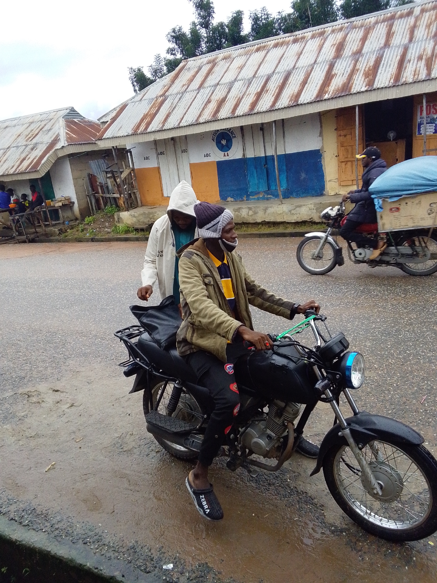 In Serti, Nigeria, a man wearing a tan jacket, a striped shirt, and a beanie is seated on a black motorcycle. He is holding the handlebars, and a green cord is wrapped around his right hand. Beside him, a person in a white hooded jacket stands, appearing to speak to the rider. The motorcycle has a black seat, a metal rack at the back, and chrome spokes on its wheels. The road is wet and appears to be paved with asphalt and gravel. In the background, buildings with corrugated metal roofs and painted walls are visible. Another person is riding a motorcycle in the background, carrying a large blue tarp-covered object on the back. The sky is overcast.