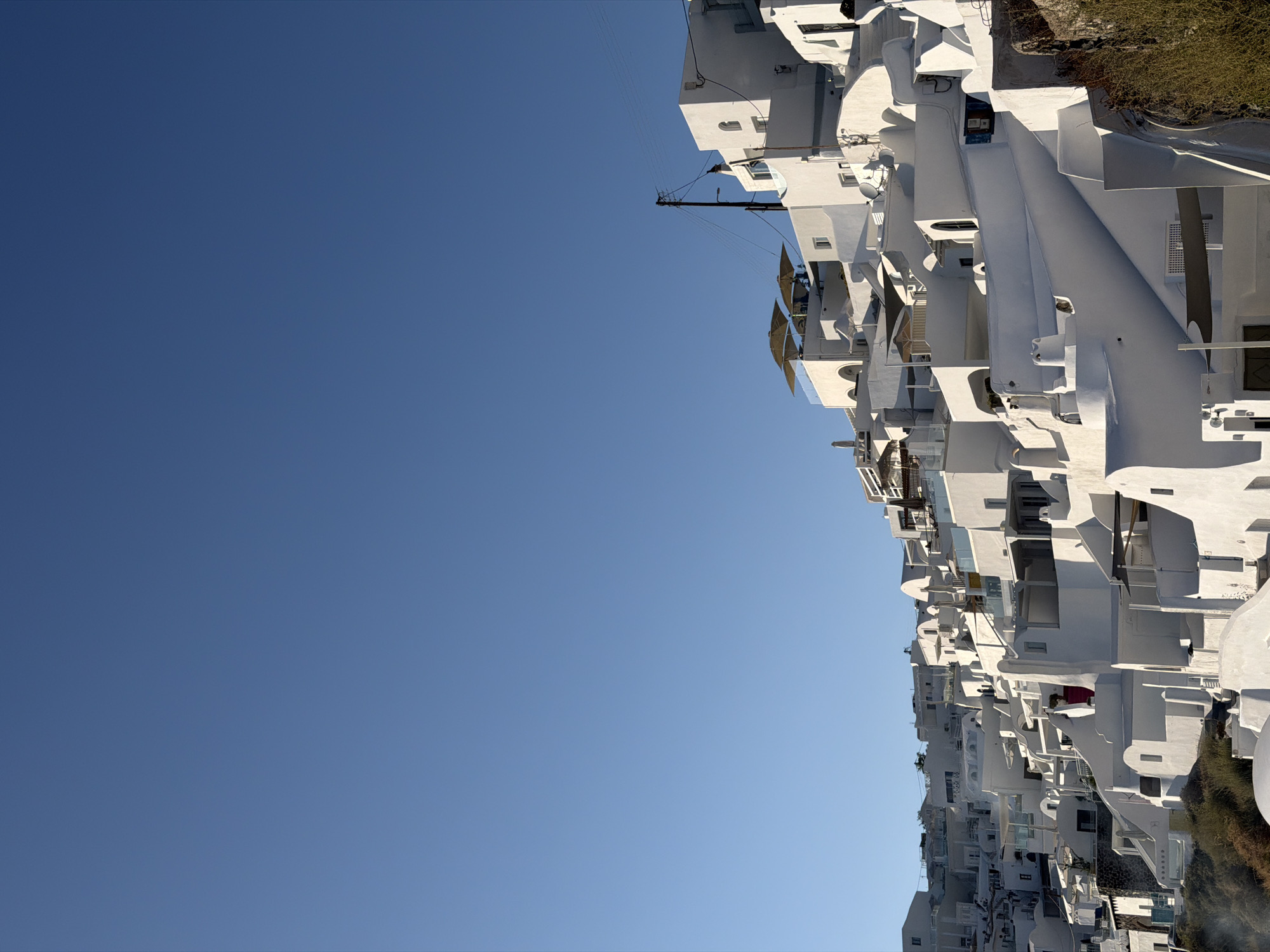 A cluster of white buildings, characteristic of Santorini architecture, cascades down a hillside under a clear blue sky. The structures are tightly packed, with varying levels, curves, and open terraces. Some terraces are equipped with umbrellas, suggesting outdoor seating or dining areas. Architectural features include arched windows, balconies, and a prominent red door on one of the buildings. Utility poles and wires are visible in the upper portion of the image. The foreground shows dry vegetation. The scene is located in the Municipal Unit of Thira, Greece, a region known for its distinctive whitewashed villages perched on cliffs overlooking the Aegean Sea. There are no people or discernible actions in the image.