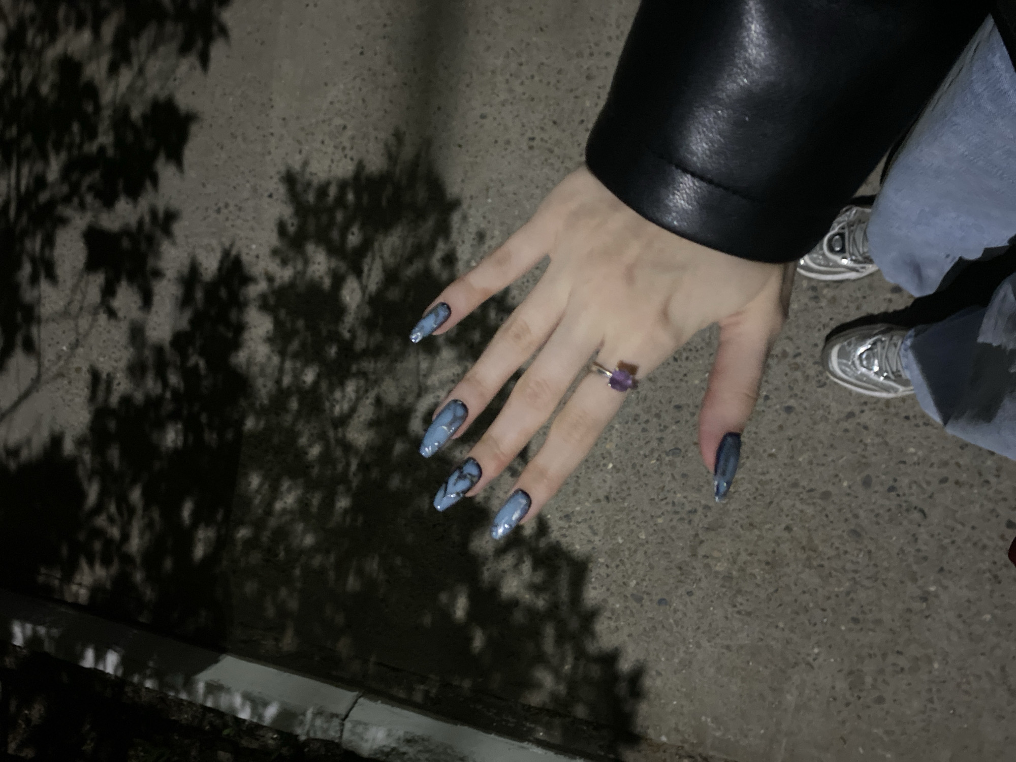 A photograph taken at night shows a person's hand with long, painted nails adorned with a blue and black marble design. The hand wears a ring with a purple stone. The person is wearing a black leather jacket and light blue jeans. In the lower-right corner of the image, a pair of silver sneakers are visible. The hand is positioned above concrete with shadows cast by foliage in the left foreground. The location appears to be an outdoor urban setting with street-level lighting.