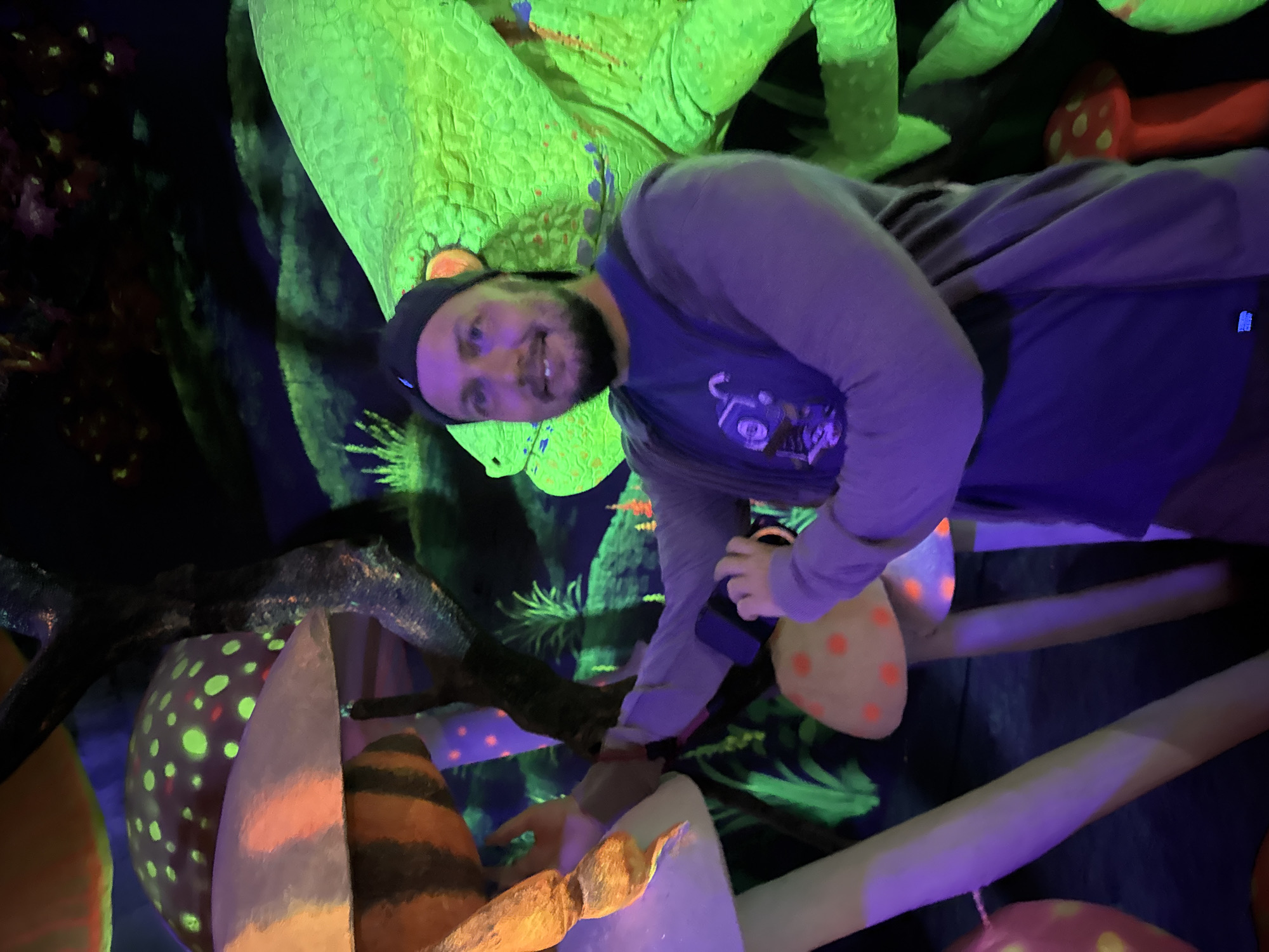 A man stands among neon-illuminated decorations in a dimly lit indoor setting. He wears a dark cap, a blue T-shirt with a graphic, and a gray long-sleeved shirt. He has a beard and is smiling towards the camera. The environment includes oversized, stylized mushrooms with colorful spots and stripes. There is also a large green dinosaur decoration in the background, seemingly painted with luminescent paint. The location in the city of Binh Lu, Vietnam, cannot be verified from the image alone. The man appears to be interacting with a mushroom-shaped object, possibly touching or adjusting it. The image has a noticeable purple tint, likely from the UV lighting.