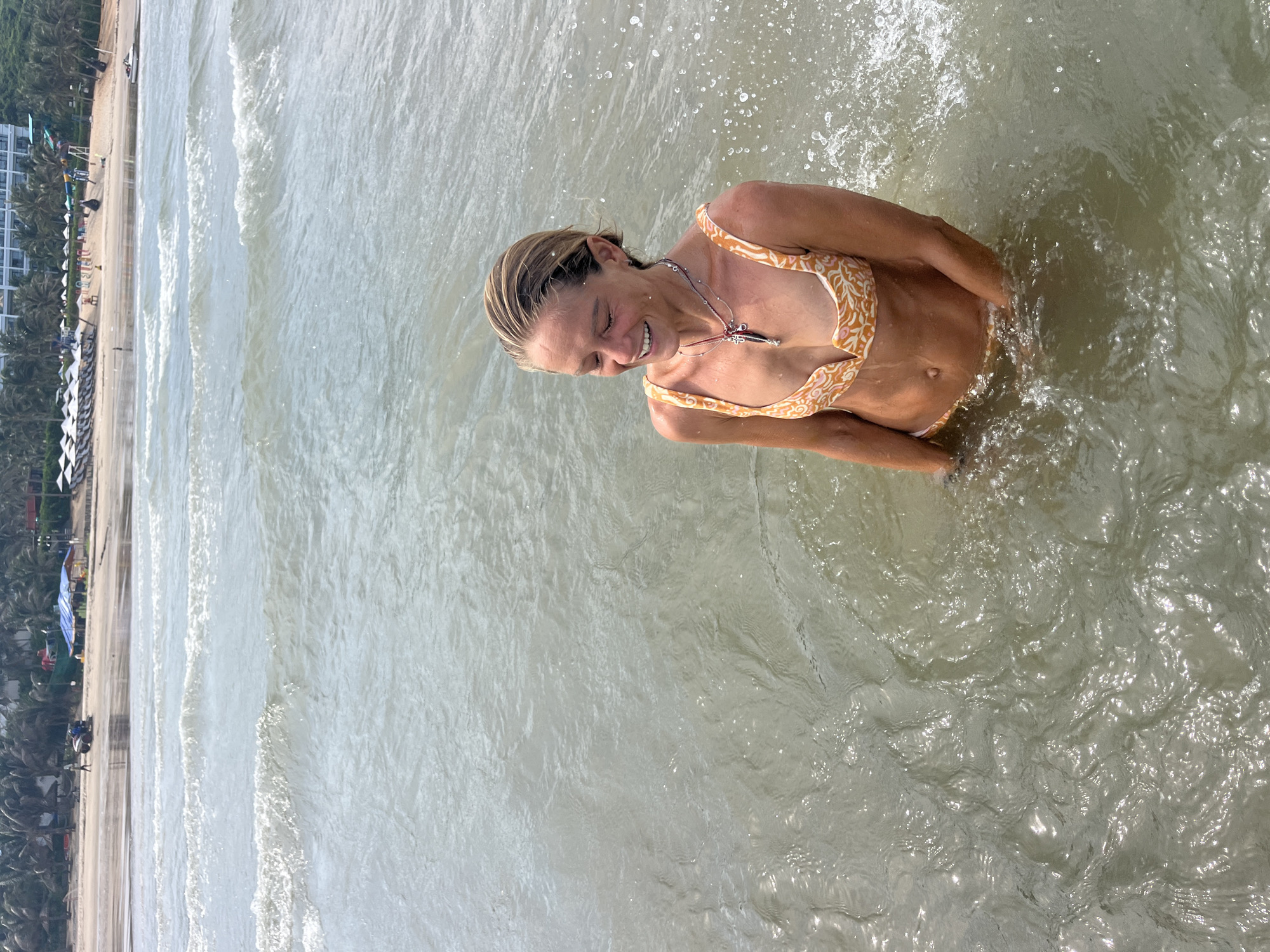 In Mũi Né, Vietnam, a woman with blonde hair, wearing an orange and white patterned bikini, stands in the ocean. She is smiling and looking down, with water reaching her waist. Her arms are submerged in the water, and splashes are visible around her. In the background, waves roll towards the shore, where a sandy beach is lined with palm trees, umbrellas, and buildings, including a resort with a water slide.