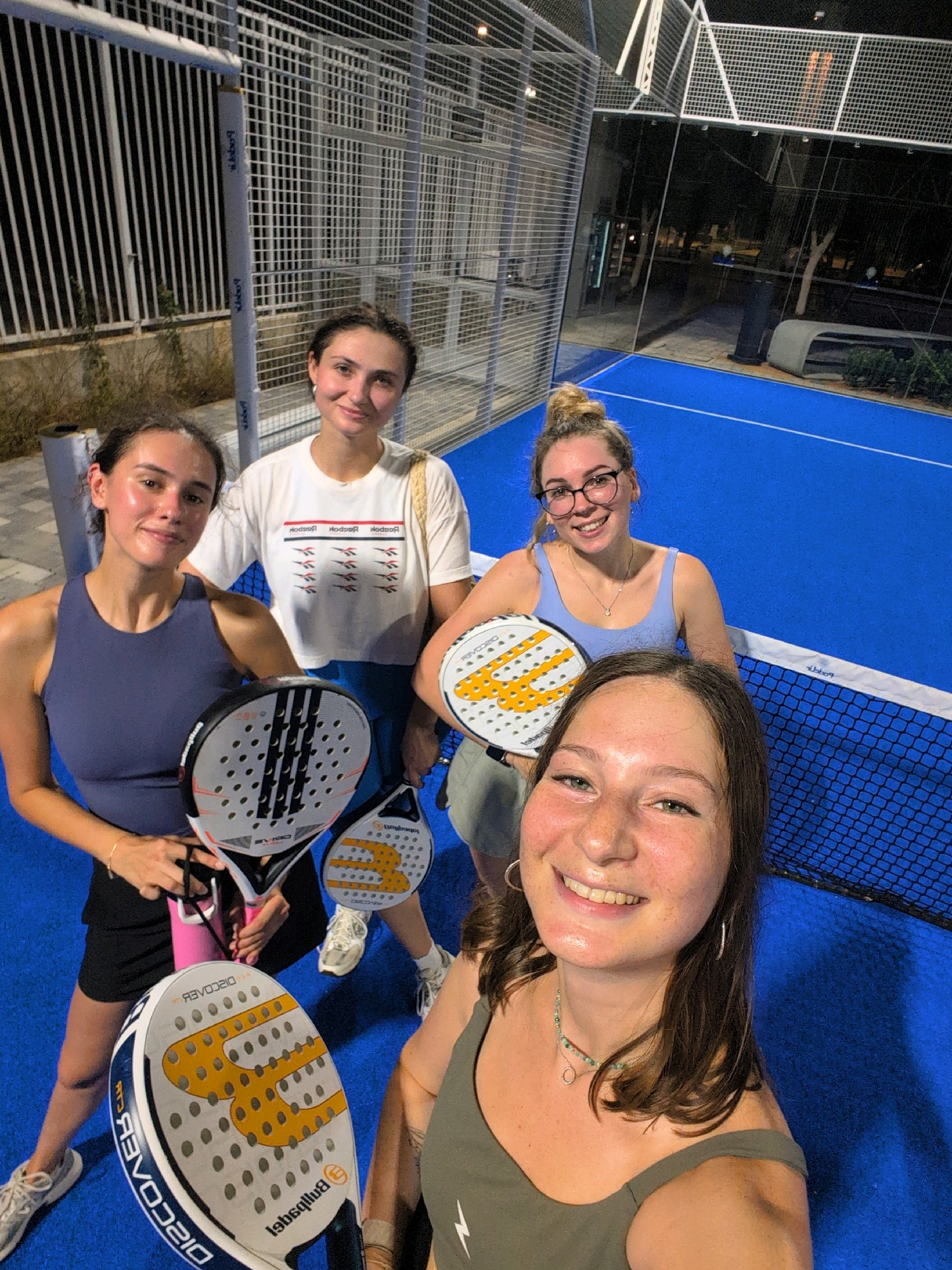 Four young women are standing on a blue padel court at night in Ramat Gan, Israel. The court is enclosed by a metal fence and has overhead lighting. The women are holding padel rackets and are smiling at the camera. One woman in the foreground is taking a selfie. Another woman in the background is wearing a white t-shirt with a Reebok logo. The court appears to be part of a sports complex.