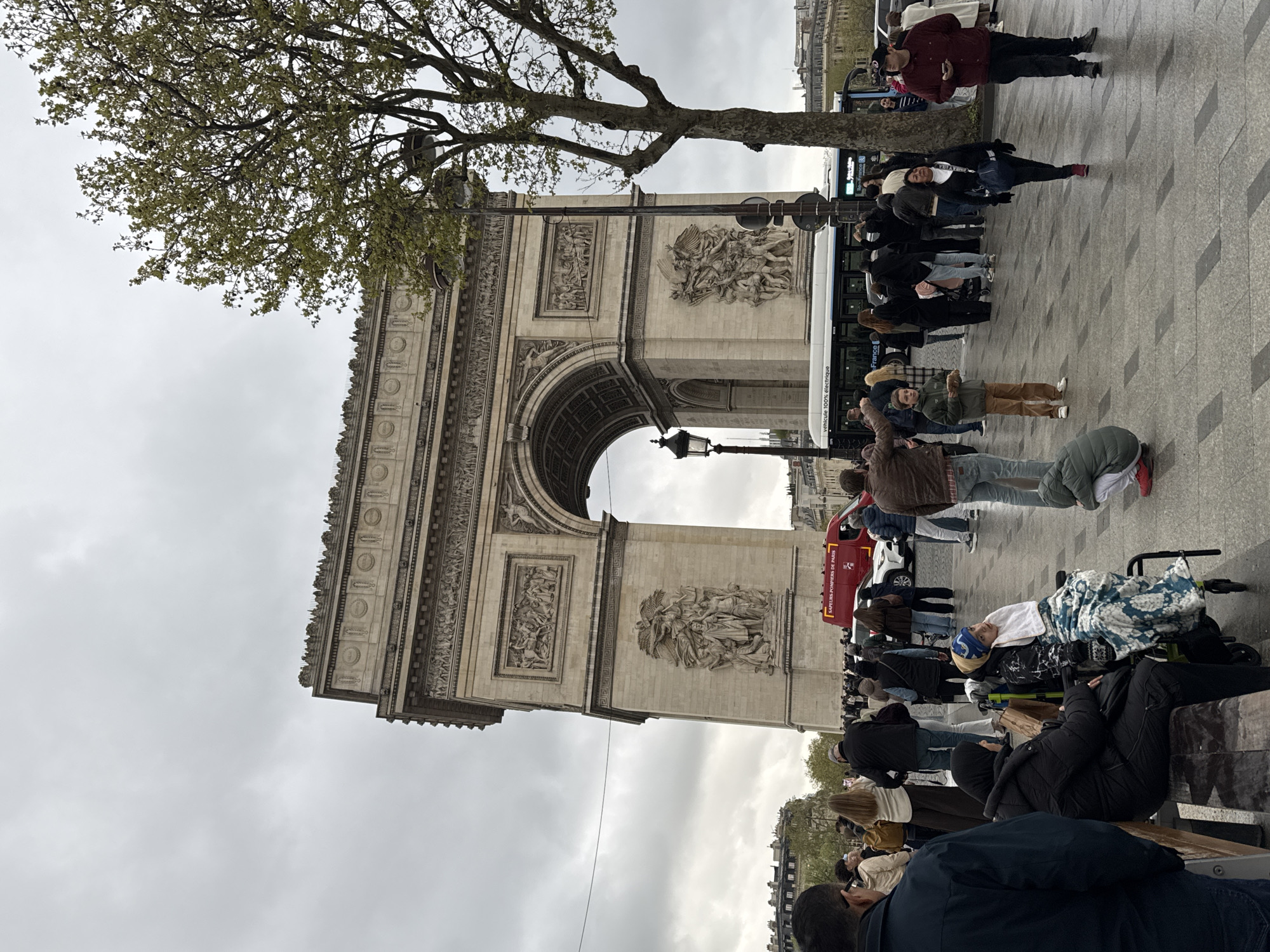 The image depicts the Arc de Triomphe in Paris, France, surrounded by tourists and city life. The sky is overcast. A tree with budding leaves is on the left side of the frame. The Arc de Triomphe is the focal point, showing its detailed carvings. People are walking around and gathering in front of the monument. There's a white tour bus parked along the street next to the monument. A red "SAPEURS-POMPIERS DE PARIS" vehicle is behind a group of people. A person is lying on a portable bed. Some people are sitting on benches along the street.
