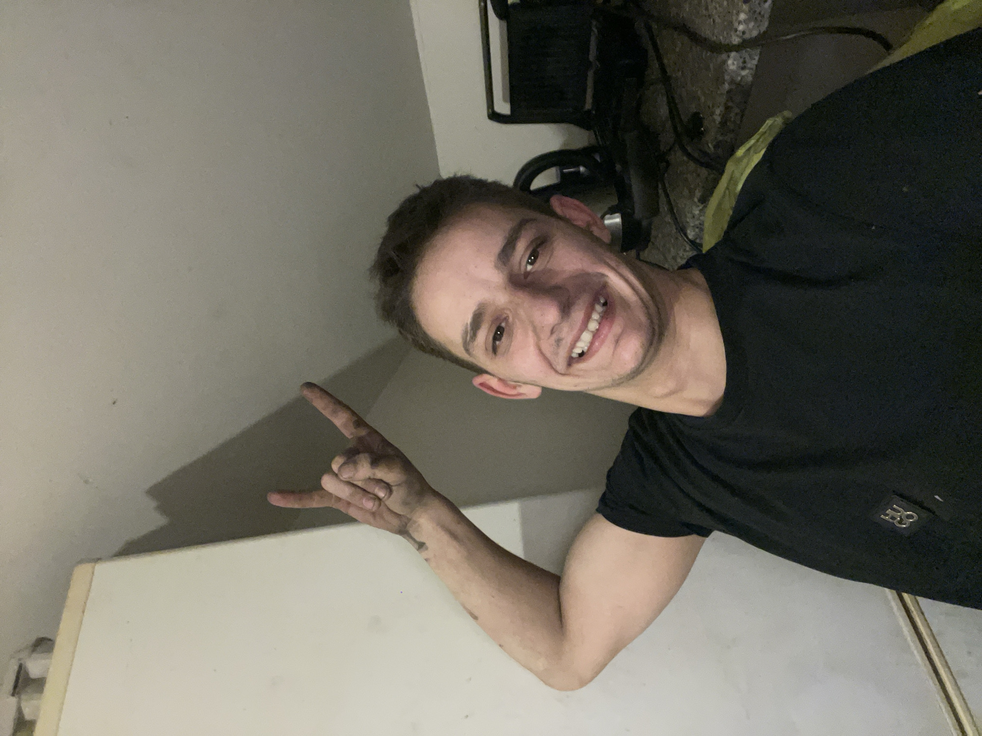 An indoor, eye-level photograph depicts a smiling young adult male in what appears to be a kitchen or utility area. The individual, with short dark hair and light skin, is positioned slightly right of center, looking directly at the camera with a wide smile revealing teeth. He is wearing a black short-sleeved t-shirt with a small, metallic-colored logo on the left chest. His right arm is raised, with his hand forming a "rock on" gesture (index finger and pinky extended). The right hand and forearm are visibly soiled with dark residue. To the left, a light-colored, upright appliance or cabinet is partially visible. To the right and behind the subject, a dark, speckled countertop holds various appliances, including a dark grill or sandwich maker and cables. The walls are a light neutral color, and shadows are cast on the wall behind the subject. The location is an unspecified room within a private dwelling in Unknown, Unknown.