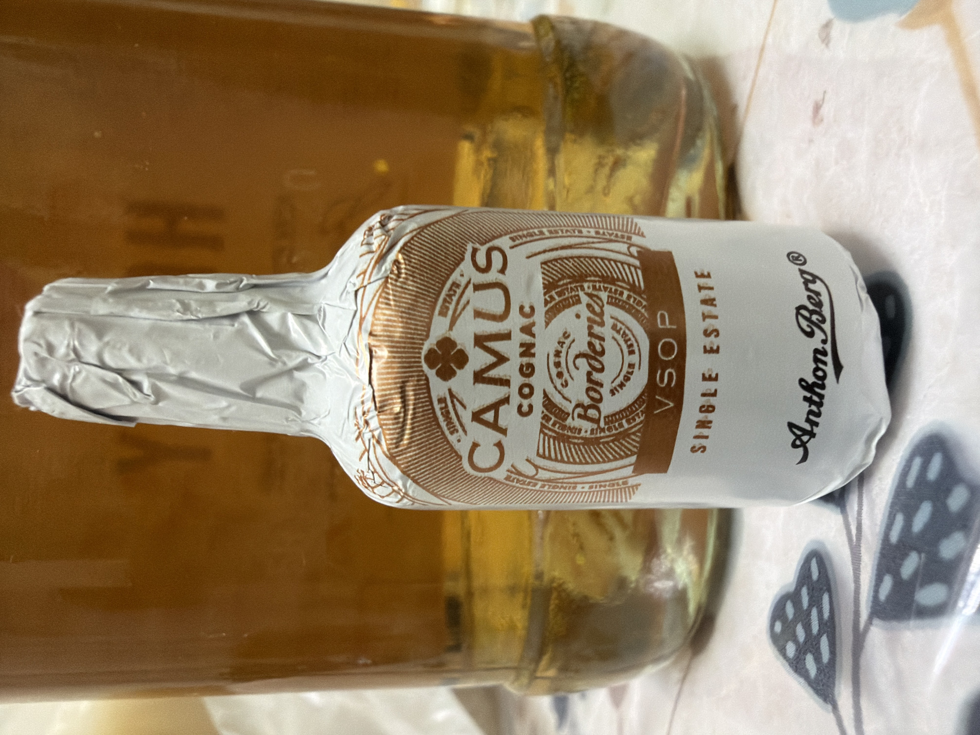 The image displays a miniature bottle of Camus Borderies VSOP Cognac, wrapped in silver foil, standing on a table. The bottle is positioned in the foreground, slightly off-center to the right, with a blurred background showing a larger glass container filled with a golden liquid, likely more of the same beverage. The table surface is covered with a patterned cloth. The lighting suggests an indoor setting. No specific location in Kyiv, Ukraine, can be identified from the image.