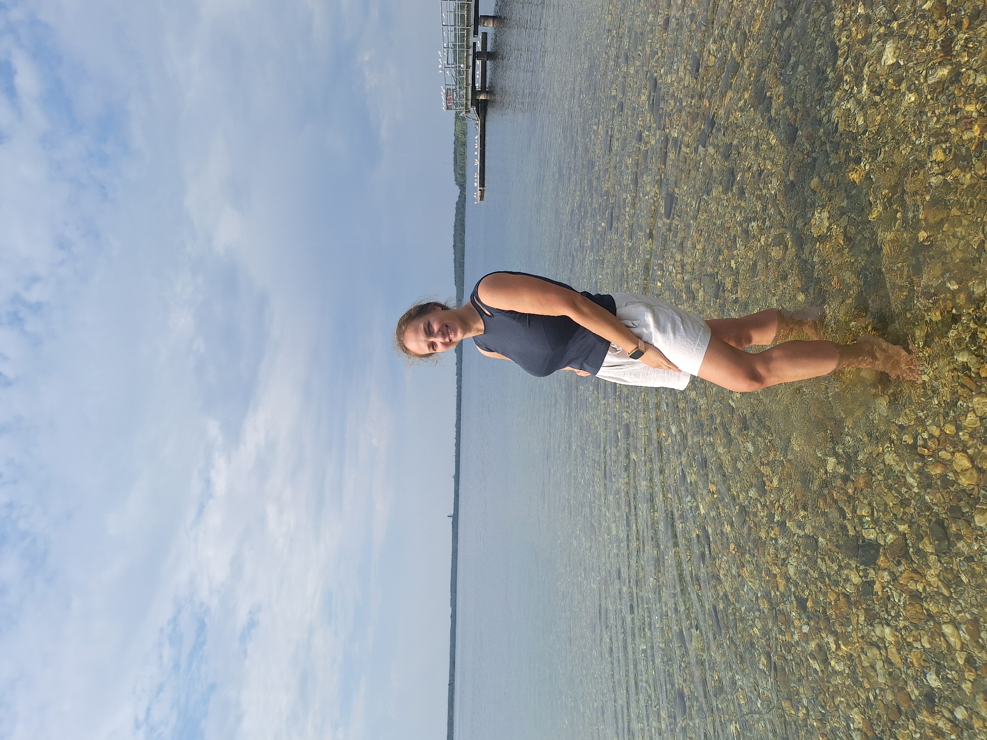 A young woman stands in shallow, clear water near the shore of a lake. She is smiling and looking at the camera, with her left hand on her hip. She is wearing a dark blue tank top and light-colored shorts. The water is clear enough to see the rocky bottom, and ripples are visible on the surface. In the background, a wooden pier extends into the lake, with several birds perched on it. The sky is partly cloudy. The scene is likely located at the Cospudener See, a popular recreational area south of Leipzig, Germany.