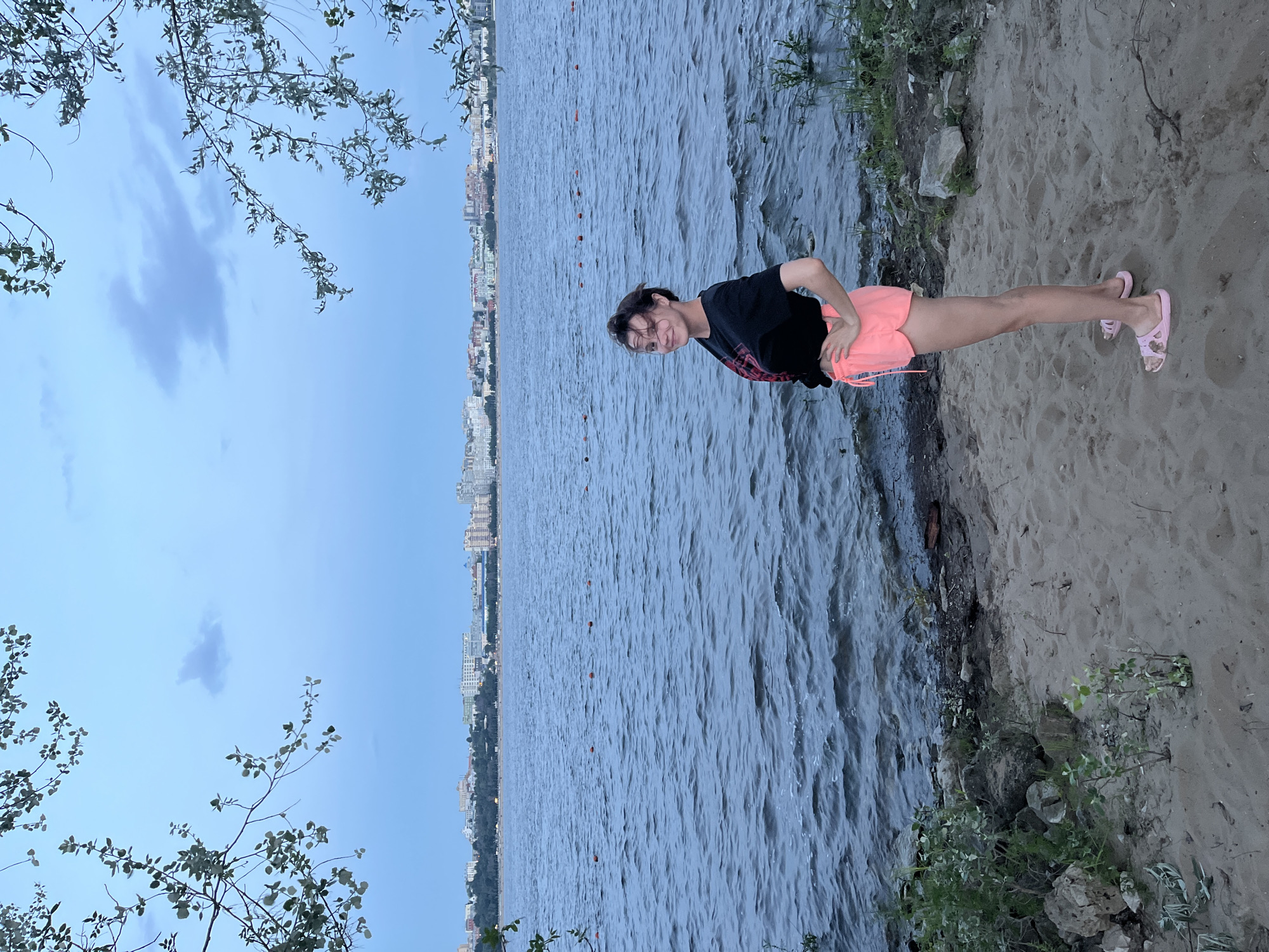 A full-shot, eye-level outdoor photograph shows a young woman standing on a sandy shore facing a large body of water. The woman is positioned in the center-right of the frame, facing the viewer with her left side slightly forward and her right hand on her hip. She has dark hair pulled back into a ponytail, is wearing a black t-shirt with a red graphic design on the front, and neon pink shorts. She has on light pink slip-on sandals.
The background features a wide, rippling body of water with a faint line of orange buoys stretching across it. In the distance, a city skyline is visible, with numerous buildings of varying heights and colors. The sky above is a dusky blue with scattered clouds. Lush green foliage drapes down from the top left and right corners of the image, framing the scene.
The shore in the foreground is composed of sand with scattered rocks and patches of green vegetation. The lighting suggests it is late afternoon or early evening. The location is identified as a shoreline in Novokuybyshevsk, Russia.