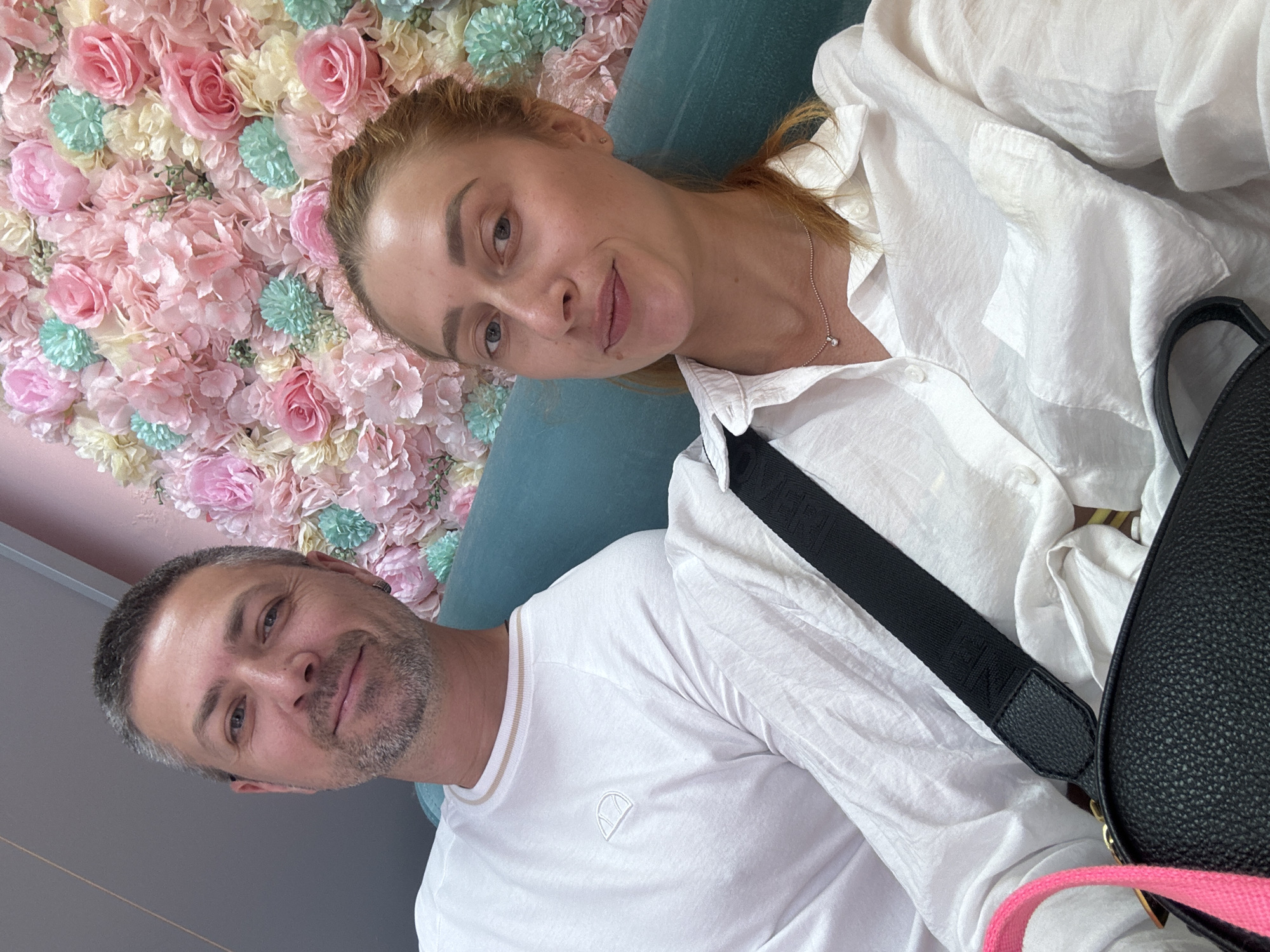 The image shows a couple sitting on a teal sofa in front of a wall covered with pink, yellow, and mint green artificial flowers. The man, on the left, is wearing a white long-sleeved shirt. The woman, on the right, is wearing a white button-down shirt and a delicate silver necklace. She has a black handbag with a black strap and a bright pink strap over her shoulder. They appear to be posing for a selfie. The background suggests an indoor setting, possibly a cafe or a photo studio, commonly found in tourist areas like Budva, Montenegro. The lighting is bright, suggesting it might be daytime.