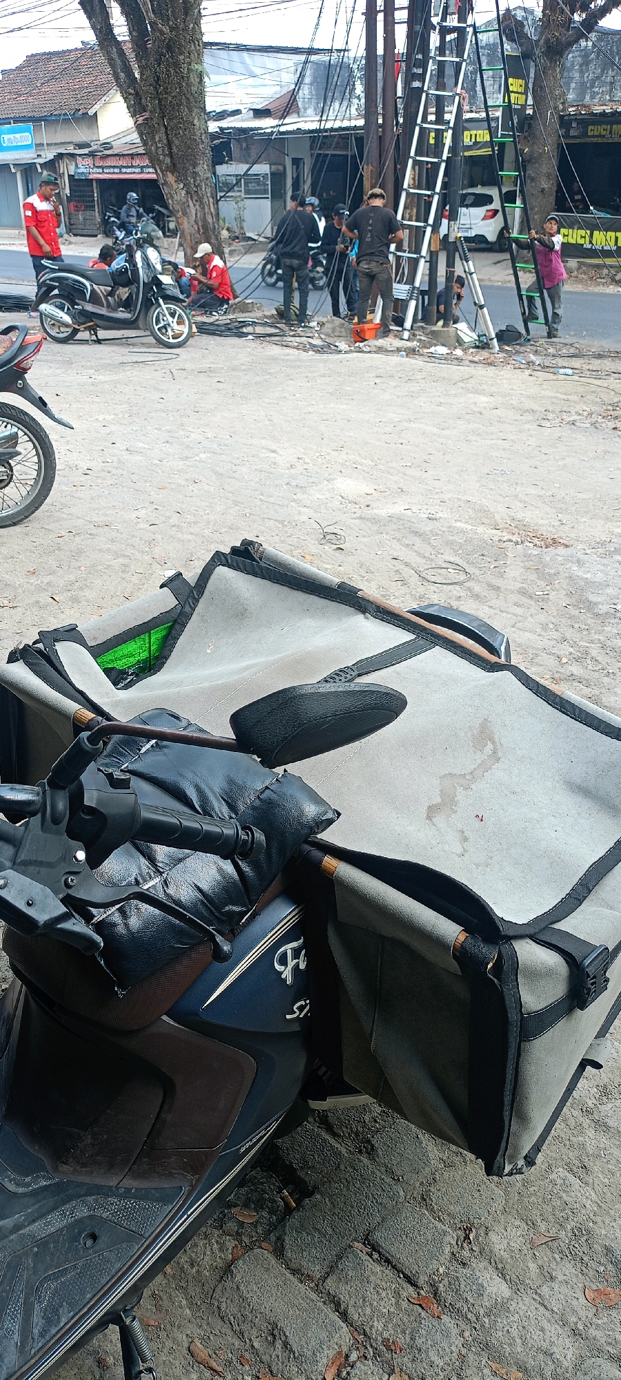 The image depicts an outdoor scene on a street in Serangmekar, Indonesia, where workers are engaged in electrical work near a utility pole. In the foreground, a scooter is parked with a large, open bag draped over its seat and handlebars. The bag appears to be made of a light-colored canvas material with black trim. The scooter has a dark blue body with some brown accents and features a padded black cover over the handlebars.
In the background, a group of individuals are gathered around a utility pole laden with numerous electrical wires. One person is standing on a tall, silver ladder, seemingly working on the wires. Others are positioned on the ground, with some appearing to be assisting or observing. A motorcycle is parked near the utility pole. The environment suggests an urban or semi-urban setting, with buildings and a road visible. The ground is a mix of dirt and paved surfaces. The overall impression is one of ongoing maintenance or repair work on the electrical infrastructure.