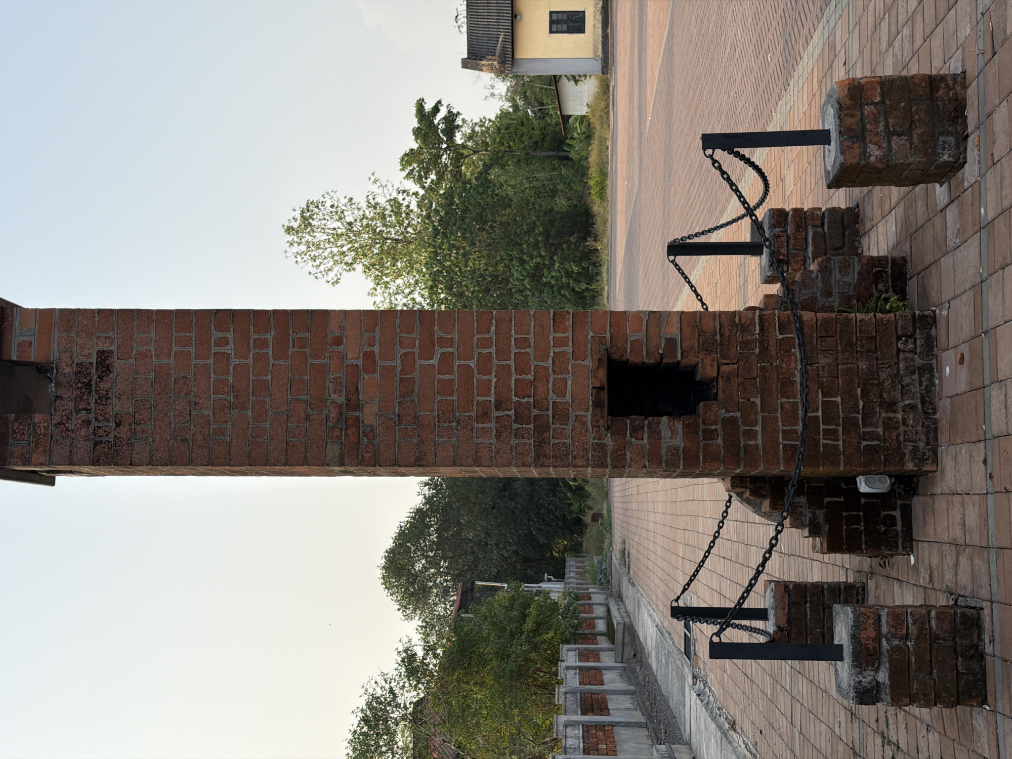 The image shows a tall, thick brick structure bisecting the frame, seemingly a section of a wall or a gateway. A rectangular opening is visible in the middle. Black metal posts with chains are attached to the structure on the right side. Beyond the structure, a brick-paved path and vegetation, including trees, are visible. In the distance, a small yellow building with a dark window can be seen. The sky is light blue and cloudless. The location is Kalamassery, India.