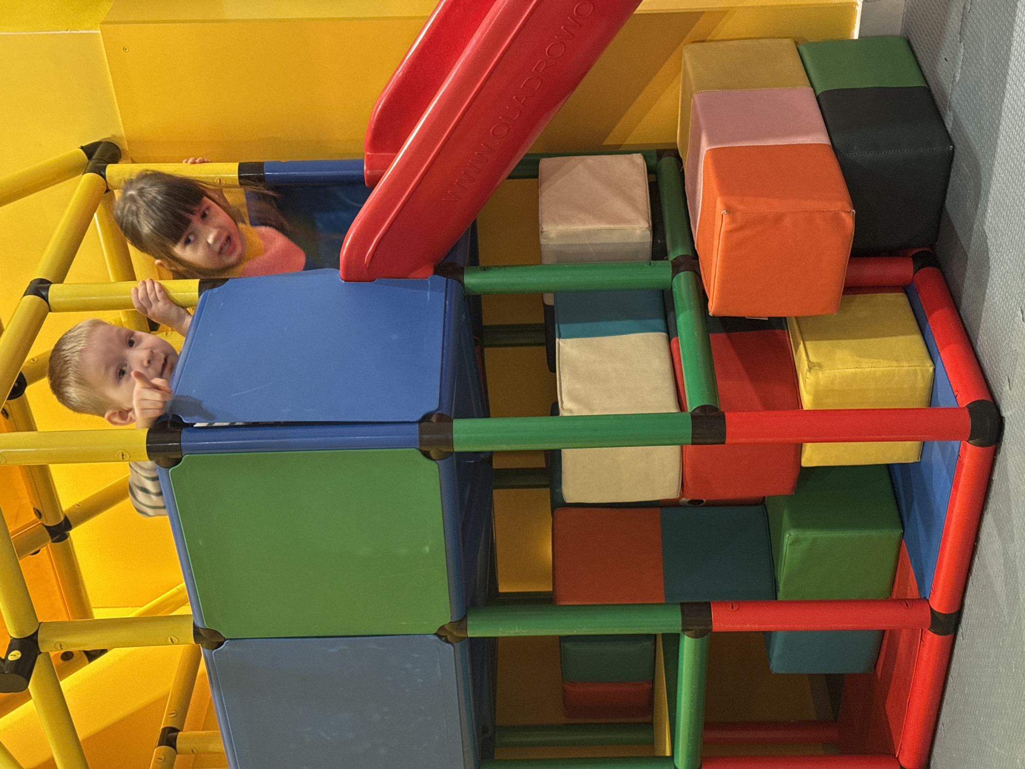 This scene is located within an indoor children's play area in Izhevsk, Russia. Two children, a fair-haired boy and a dark-haired girl, are visible from the chest up, looking out from an elevated platform of a multi-level modular play structure. The boy, on the left, wears a striped shirt and points forward with his right index finger, exhibiting a slight smile. The girl, on the right, has bangs, wears a pink and orange top, and looks forward with a neutral expression while gripping a blue panel of the structure.
The play structure is constructed from interlocking plastic modules and support poles in bright colors, including blue, green, red, and yellow. The upper framework, above the children, is yellow. A red plastic slide, partially inscribed with "WWW.QUADROWO", descends from the children's platform on the right. Below the children's level, numerous soft, upholstered blocks in various colors (red, blue, green, yellow, orange, pink, cream, dark grey) are arranged within and around the lower sections of the play structure. The floor visible is a textured grey safety mat. The background walls are a uniform, bright yellow, indicating an artificially lit interior.