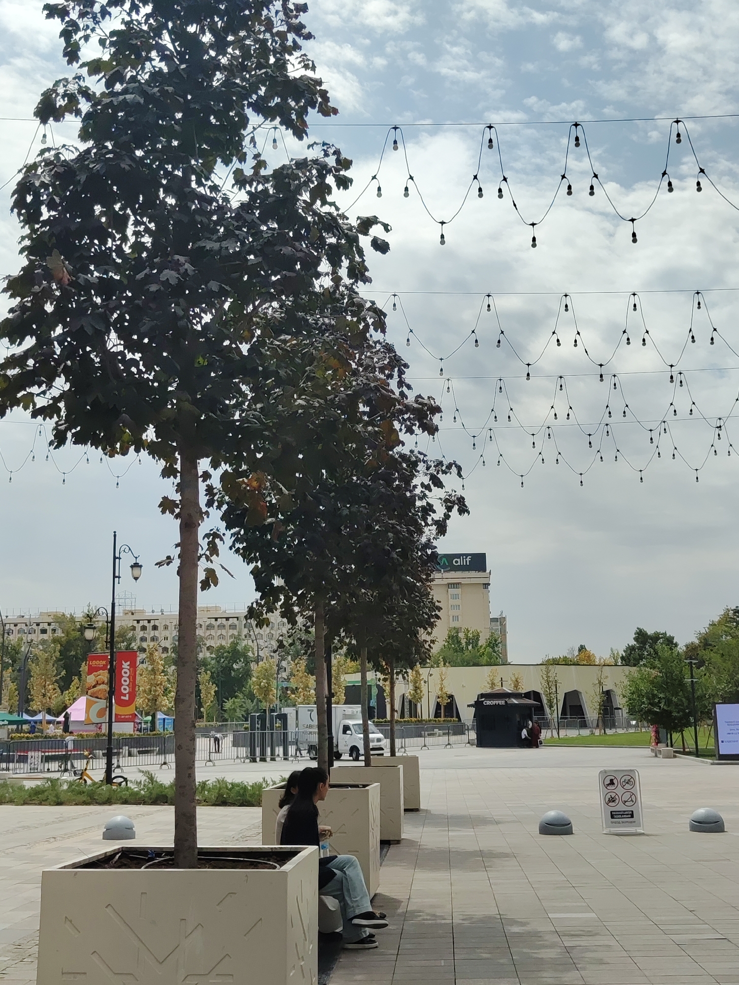 In Tashkent, Uzbekistan, an outdoor scene unfolds on a paved plaza under a cloudy sky. Strands of decorative light bulbs are strung across the sky in an undulating pattern. In the foreground, a person sits on a bench, partially obscured by a large planter box containing a dark-leaved tree. The person is wearing a black top and blue jeans, with their legs crossed and a water bottle in their hand. To the right of the bench, another planter box is visible.
Further back, a small coffee kiosk with the word "CROFFEE" is visible. Several other trees are planted in planters across the plaza. In the mid-ground, a white van is parked. A red banner with "LOOOK" printed on it hangs from a lamppost. In the background, a multi-story building with many windows stands prominently, and a sign with "ALIF" is visible on its facade. Additional trees and market stalls can be seen in the distance, suggesting a lively public space. A few round, grey bollards are scattered across the plaza. A sign with various icons, including those prohibiting pets, bicycles, and smoking, is placed on the pavement. The overall impression is a modern urban setting with elements of leisure and commerce.