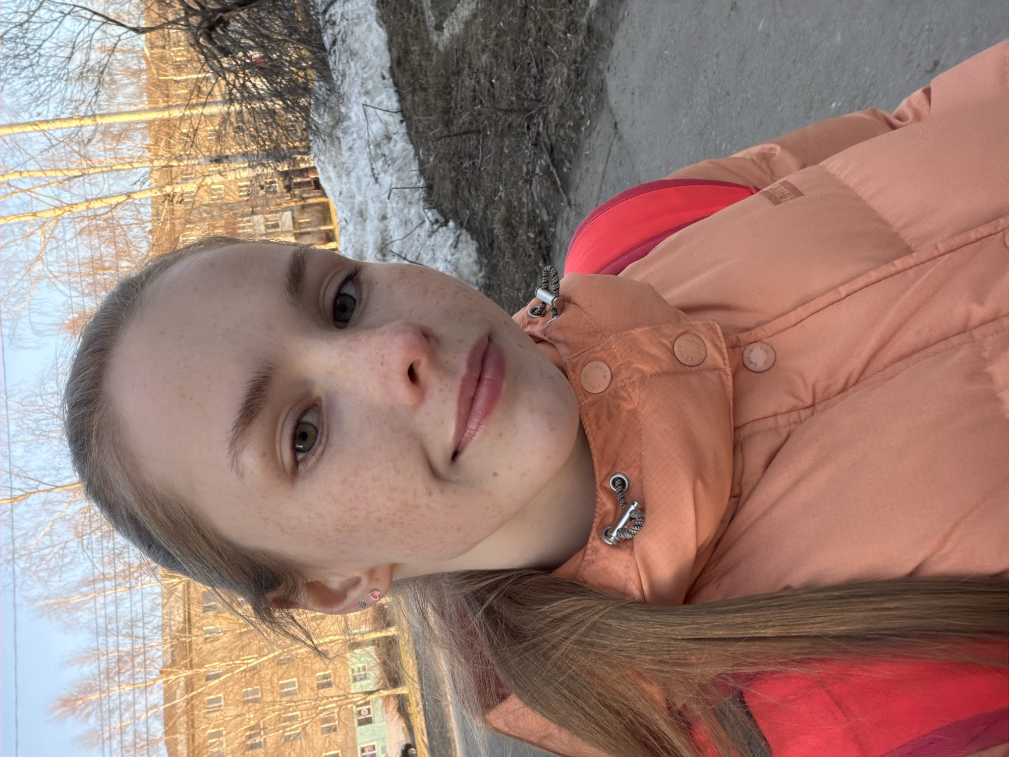 A young, fair-skinned woman with freckles is posing for a selfie. She has light brown hair and is wearing a peach-colored puffer jacket with red accents. The background appears to be an urban environment, likely in Berezniki, Russia, given the bare trees, patches of snow, and indistinct building visible. The photo seems to have been taken during the day.