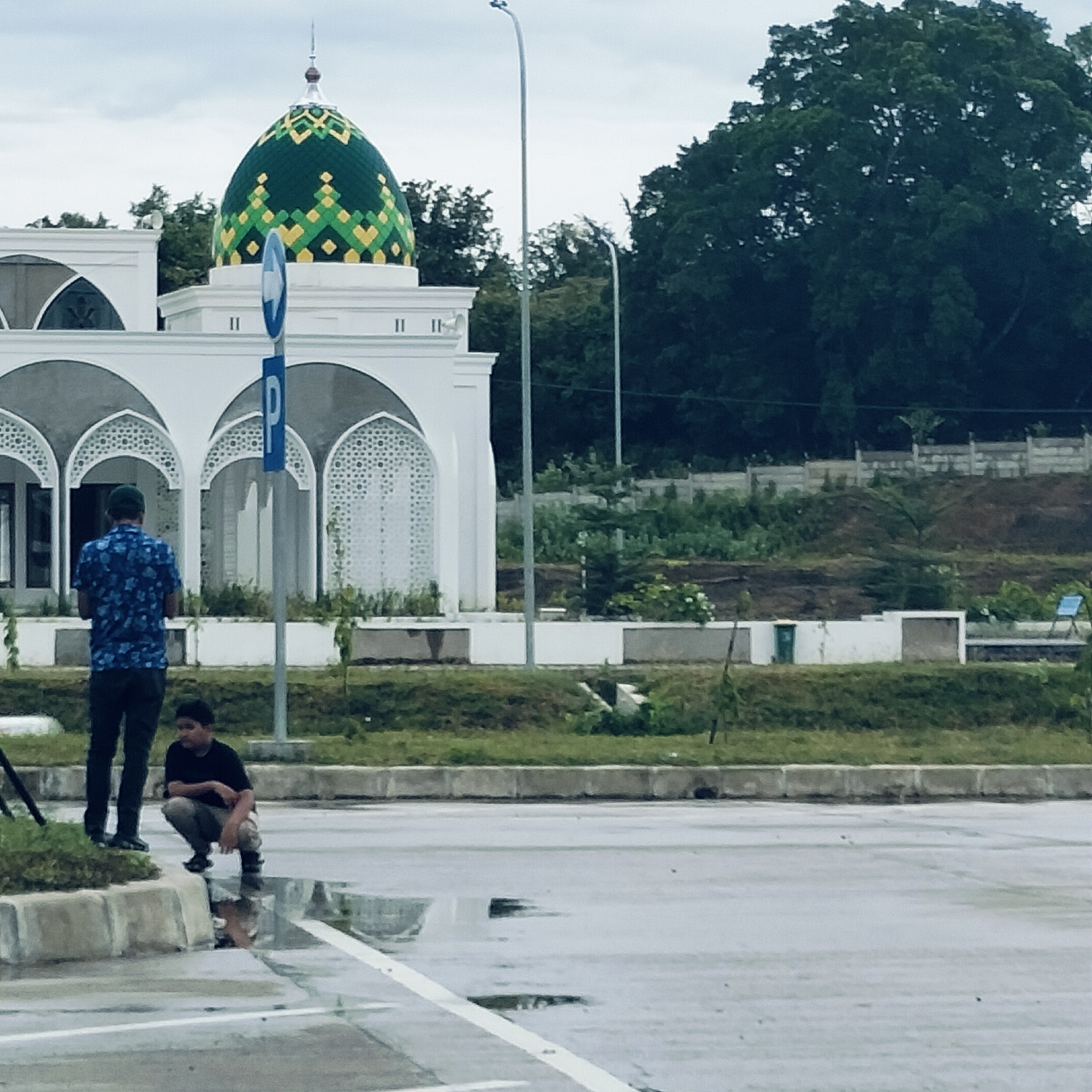 The image captures an outdoor scene featuring a mosque, two individuals, and surrounding elements under an overcast sky, likely in Keureuweungkrueng, Indonesia.
The central subject is a white mosque building on the left, characterized by intricate arched windows and facades with decorative patterns. Its dome is a striking feature, adorned with a vibrant green and yellow geometric or mosaic design, topped by a slender, pointed finial. To the right of the mosque and behind the people, a tall, slender streetlight stands against the sky.
In the foreground, two males are visible. One man stands with his back to the viewer, wearing a patterned blue shirt and dark trousers. Beside him, a younger male squats near the edge of a paved surface, looking towards the right. The ground is wet with visible puddles, suggesting recent rain.
The immediate foreground consists of a paved area, possibly a road or parking lot, marked with white lines and bordered by concrete curbs. Beyond this, a strip of grass separates the paved area from the mosque structure. The background is dominated by lush green trees and some elevated dirt mounds with concrete barriers.
A blue rectangular sign displaying a white letter 'P' and an arrow is mounted on a pole near the mosque, indicating a parking area. The overall atmosphere is calm, with the cloudy weather lending a soft light to the scene. No specific activity or strong emotions are discernible from the individuals; they appear to be paused or observing their surroundings.
