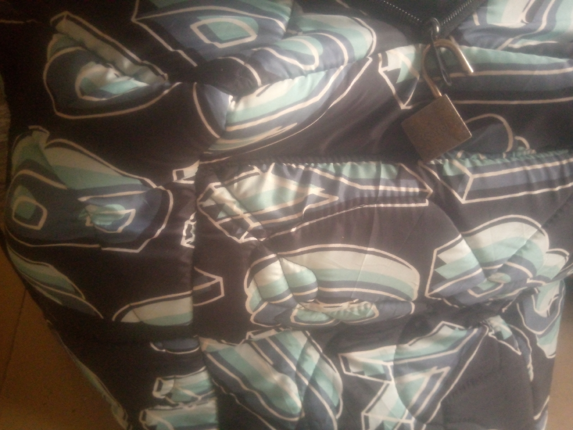 The image shows a close-up, abstract view of a dark-colored puffer jacket with a blue, white, and teal geometric pattern. A small, brass-colored padlock is attached to a zipper pull on the jacket. The lighting is dim, and the background is out of focus, suggesting the jacket is likely indoors or in a dimly lit area. There are no people or other discernible objects in the frame. The location is unknown.