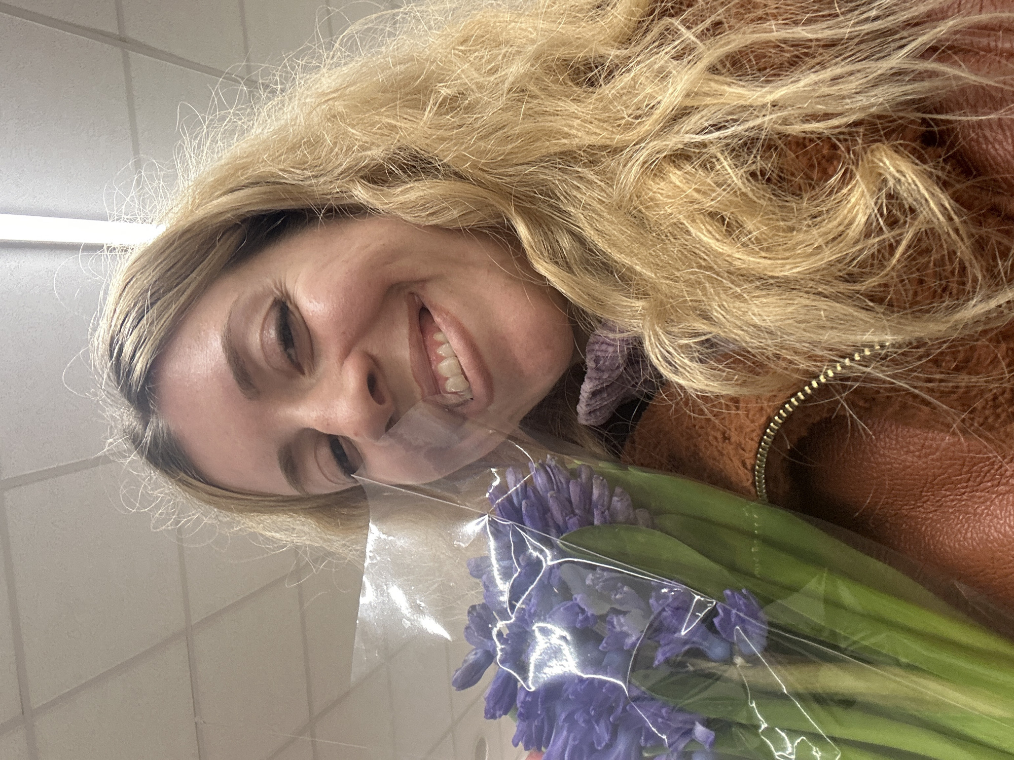 In a retail interior within the city of London, United Kingdom, a close-up, upward-angled selfie depicts a Caucasian woman. She has long, wavy blonde hair and is smiling broadly at the camera, revealing her teeth and causing her right eye to squint slightly. She wears a brown jacket with a visible brass-colored zipper along the right shoulder line and a purple top underneath. In the lower left quadrant of the frame, she holds a bouquet of vibrant purple flowers, consistent with hyacinths, encased in clear plastic wrapping. The background reveals a white suspended ceiling with rectangular acoustic tiles and integrated fluorescent strip lighting.