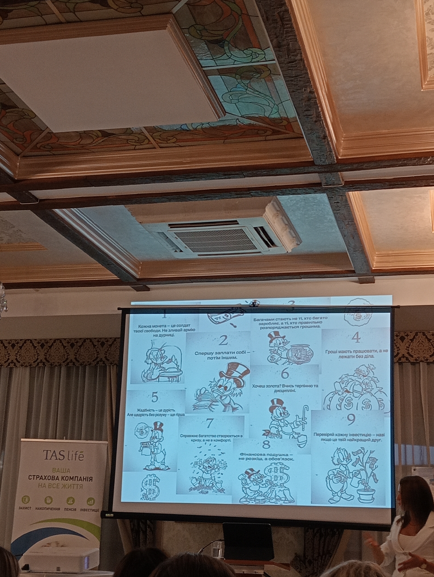 The image shows a presentation screen displaying a series of illustrated financial advice points, interspersed with cartoon characters that appear to be from a Disney production. The advice points are numbered from 1 to 9 and written in Ukrainian. The screen is positioned in a room with a patterned ceiling and wooden beams. To the left of the screen is a white banner with Ukrainian text that translates to "TAS life YOUR INSURANCE COMPANY FOR ALL LIFE" and lists "PROTECTION ACCUMULATION PENSION INVESTMENT" at the bottom. A woman in a white blazer is standing to the right of the screen, gesturing with her hands as if speaking. The audience is partially visible in the foreground, with heads and shoulders of people seated. The overall scene suggests a business or educational presentation taking place indoors. It is not possible to determine the specific location within Lviv, Ukraine from the provided image.