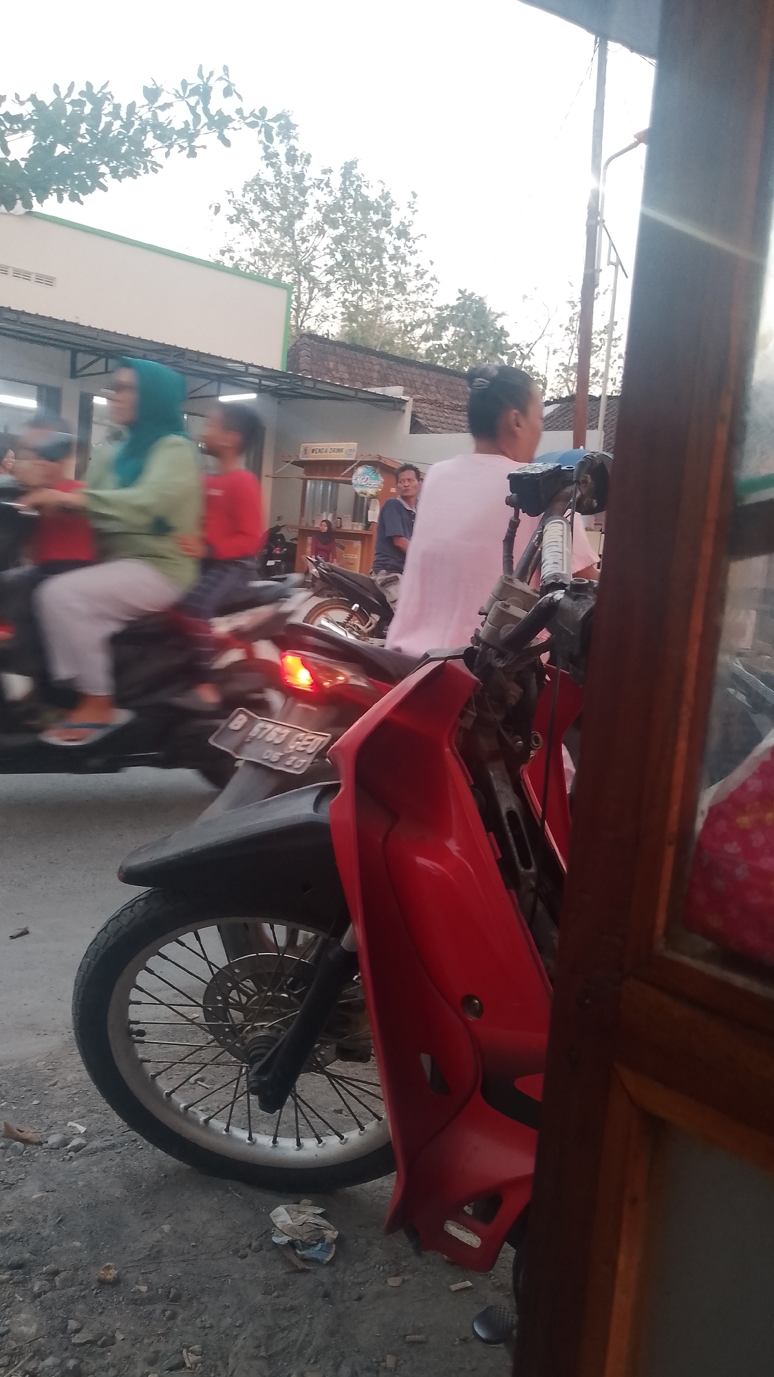 A street scene in Karanglangu, Indonesia, shows several people on and around motorcycles. In the foreground, a red motorcycle is parked, with its front wheel visible. Behind it, other motorcycles and people are blurred, indicating movement. A woman in a green hijab rides on a motorcycle, and another person in a red shirt is seated behind her. Further back, a person in a light pink garment is standing. A stall labeled "VENDA DRINK" is visible in the background. The environment appears to be an outdoor setting, possibly a roadside market or thoroughfare, with a building and trees also present. The lighting suggests it is daytime, possibly late afternoon due to the soft light.