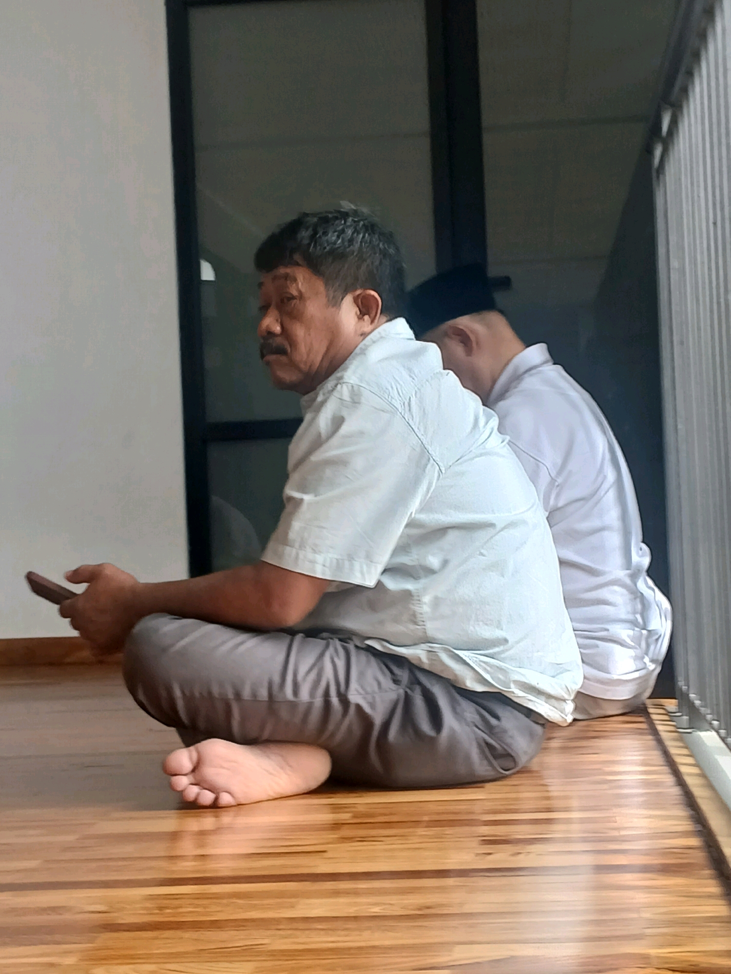 Two men are seated on a wooden floor, likely in a covered outdoor area with a railing visible to the right. The man in the foreground is looking to the left, holding a dark object, possibly a phone, in his left hand. He is wearing a light blue collared shirt and grey pants. His right leg is bent and tucked under him, with his foot visible. The man behind him is wearing a white shirt and a black cap. He is facing away from the camera, with his head bowed. The background includes a large window or glass door. The scene appears to be located in a residential setting in Arjasari, Indonesia.