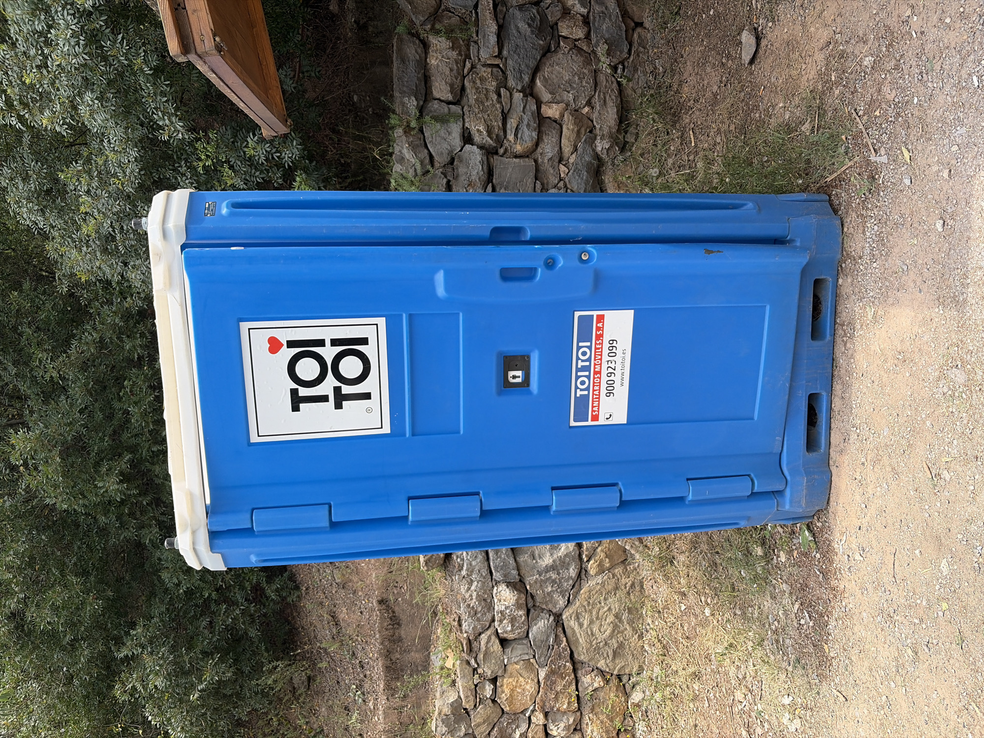 A blue plastic portable toilet, manufactured by TOI TOI, is situated outdoors in Montanejos, Spain. The toilet sits on a dirt and gravel ground surface, adjacent to a stone wall. The environment suggests a rural or semi-rural location, evidenced by the presence of uncultivated ground and the stone structure. A patch of vegetation, likely bushes or trees, is visible to the left of the frame.
