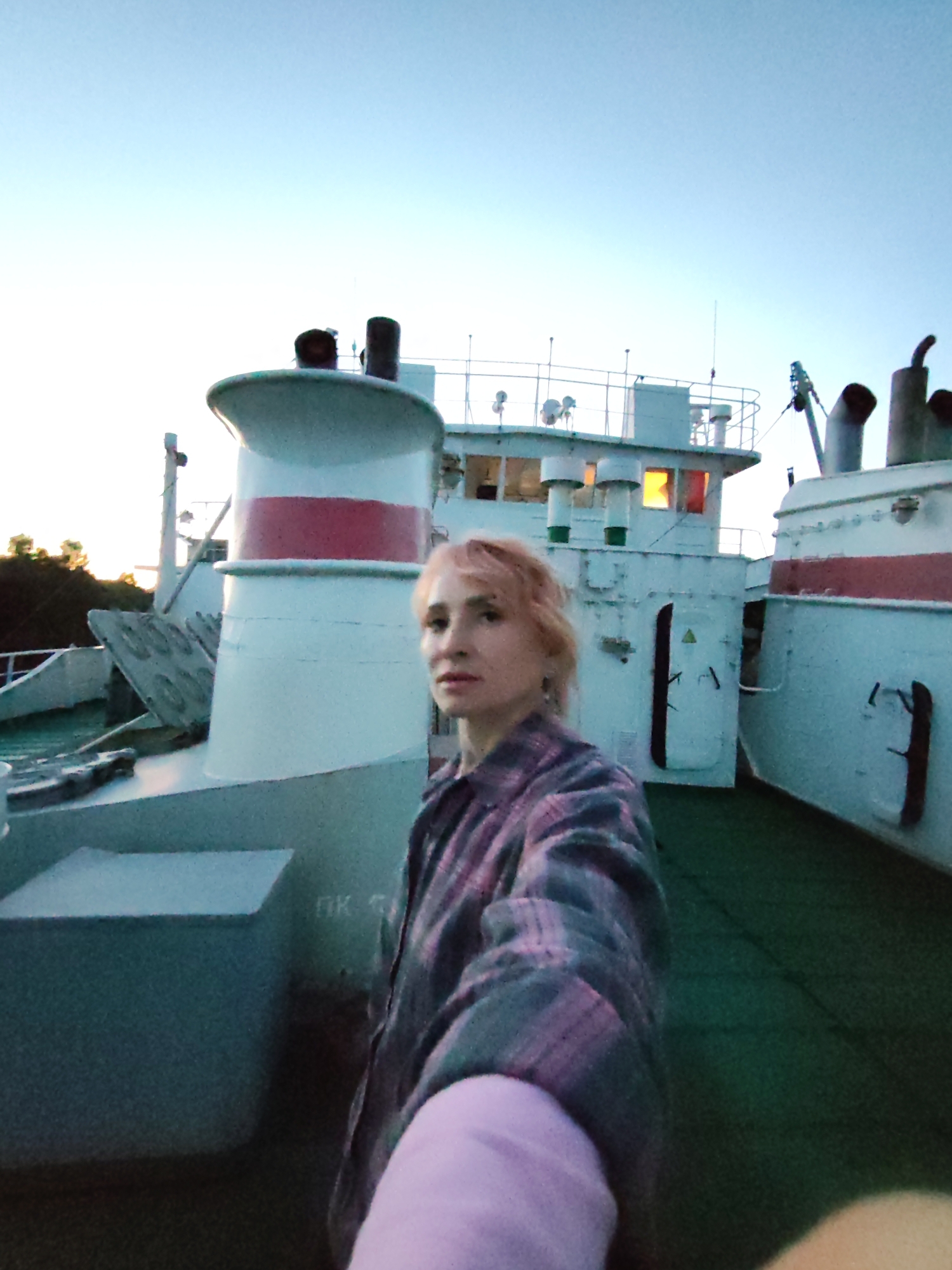 The image depicts a woman standing on the deck of a ship, with a clear sky overhead and a hint of vegetation in the background. The ship's structure, including a large white funnel with a red band and other white superstructures, is visible. The woman, with blonde hair and wearing a plaid shirt, is positioned in the foreground, looking towards the camera. The scene appears to be set at dusk or dawn, given the lighting conditions. The location is identified as Annensky Most, Russia, suggesting it could be on a river or a body of water within or near the town. The overall atmosphere is calm and serene.