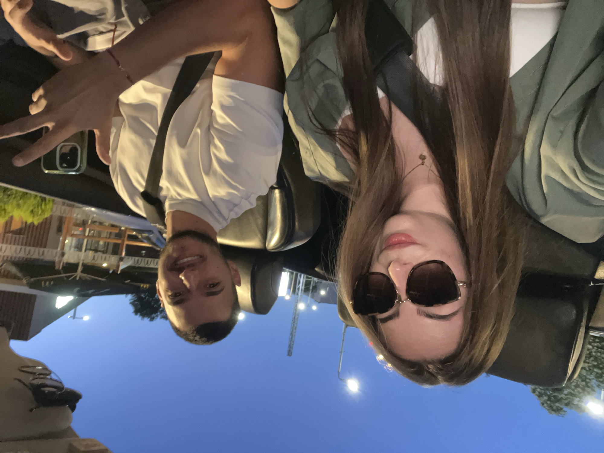 A selfie taken from inside a car at dusk in Plovdiv, Bulgaria, features a young couple seated in the front seats. The woman, on the left, has long brown hair, wears large sunglasses, a green jacket over a white top, and a delicate necklace. The man, on the right, has short dark hair and a beard, wears a white t-shirt, and is holding up two fingers in a peace sign. Both are wearing seatbelts. The car appears to be a convertible with the top down. In the background, streetlights illuminate the twilight sky, and the shapes of buildings and outdoor seating areas of a restaurant are visible. The overall atmosphere suggests a casual evening outing in the city.