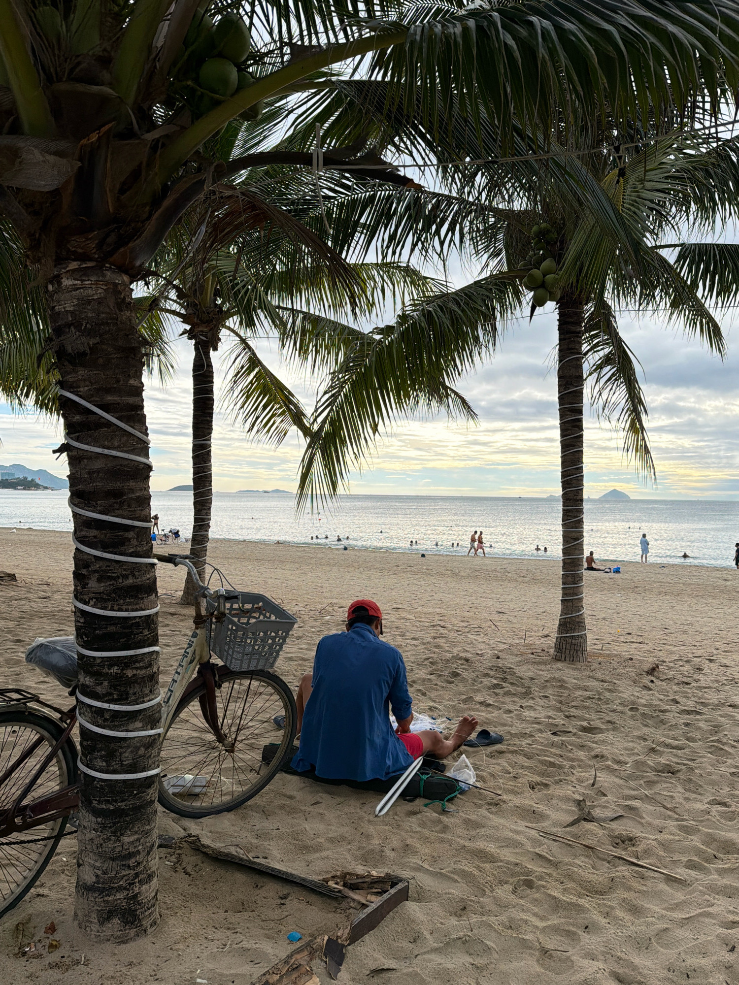 The image depicts a tranquil scene on a beach, likely in Nha Trang, Vietnam, given the presence of coconut trees and the overall coastal environment. In the foreground, a person wearing a blue shirt and red shorts is seated on the sand, facing away from the camera and towards the ocean. They are resting on a black mat with fishing gear beside them. To the left, a bicycle is parked next to a coconut tree, which is wrapped with white lights. In the background, the calm ocean stretches to the horizon, with a few people visible swimming or walking along the shoreline. The sky is overcast with soft clouds, suggesting either early morning or late afternoon. The beach itself is sandy, with sparse vegetation and debris scattered across it. The overall atmosphere is peaceful and relaxed, with a sense of solitude and connection to nature.