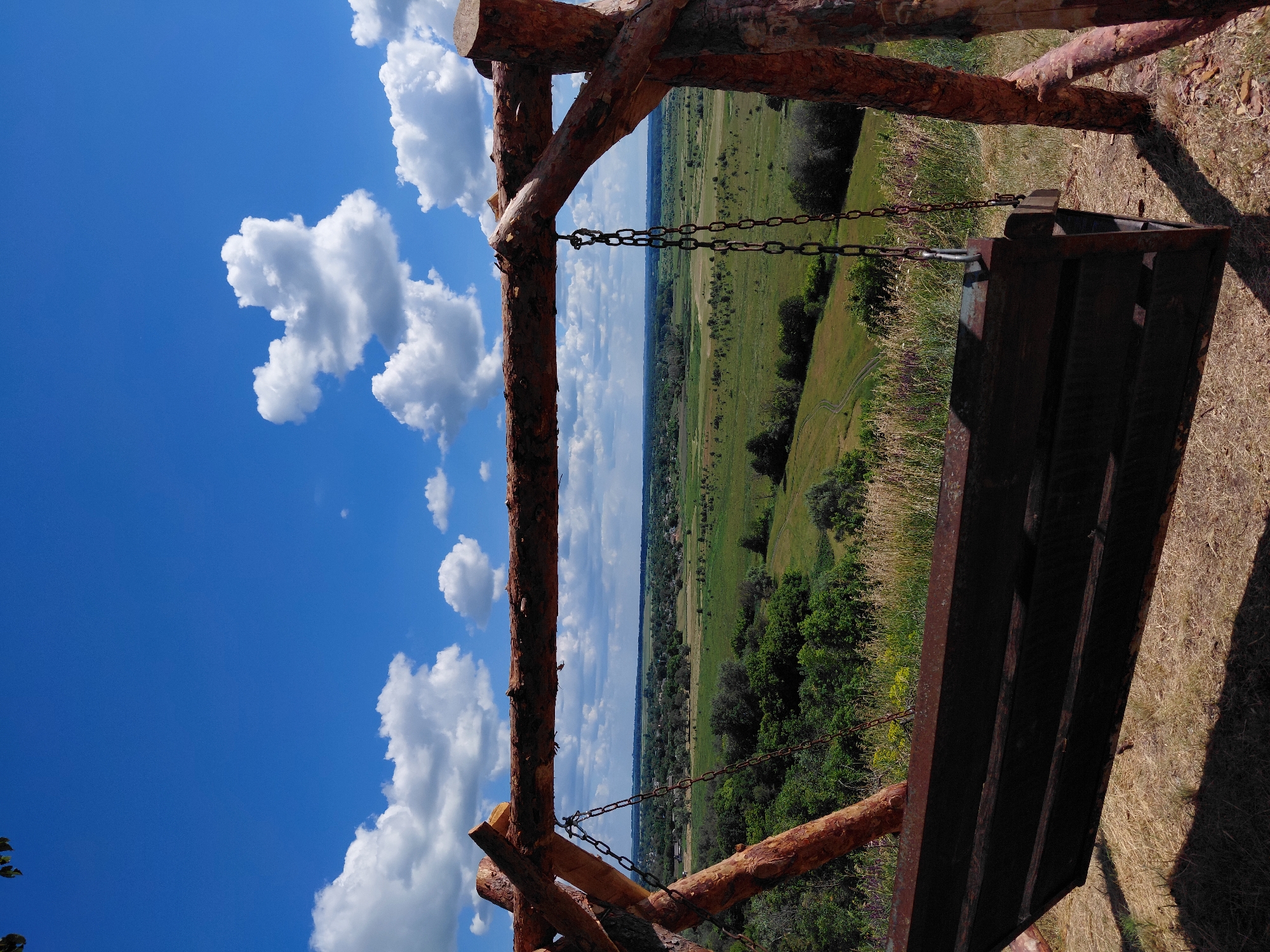 An outdoor shot captures a rustic wooden swing set overlooking a vast, green landscape under a bright blue sky filled with fluffy white clouds. The swing's bench, made of dark wood with visible slats and rusty metal edges, hangs by sturdy chains from a rough-hewn wooden frame. The frame, constructed from natural branches and logs, suggests a handmade and weathered aesthetic. The ground beneath the swing is covered in dry, light-brown grass. In the background, a rolling expanse of green fields and scattered trees stretches towards a distant horizon dotted with a few scattered buildings, possibly a village. A winding dirt path is visible in the middle ground, adding a sense of depth and leading into the landscape. The overall impression is one of a peaceful, elevated vantage point offering a panoramic view of a rural environment on a clear, sunny day.