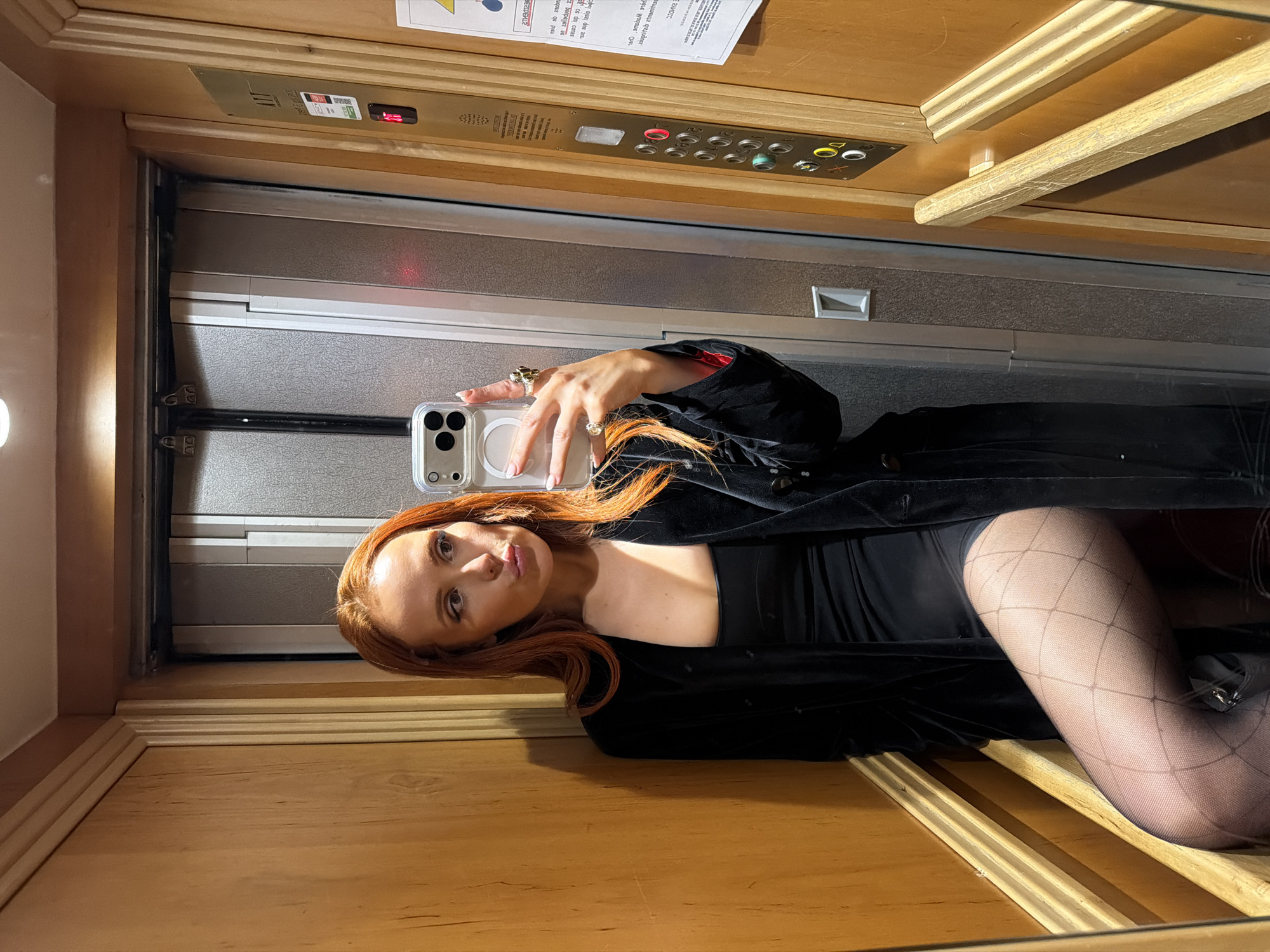 A woman with long red hair is taking a selfie in an elevator. She is wearing a black dress with a black velvet jacket and fishnet stockings. The elevator has wood paneling and a panel with buttons. A sign is attached above the elevator buttons, although the text on the sign is in a language other than English. The location of the elevator cannot be determined from the image.