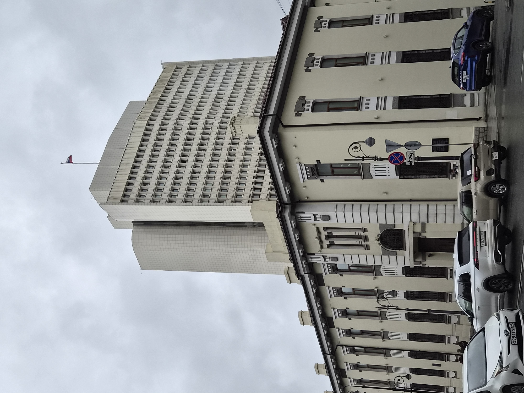 The image shows a Russian flag flying on top of a tall, modern building in Vladivostok, Russia. In front of the tall building is a shorter, older building with a light-colored facade and dark windows. Several cars are parked on the street in front of the older building, including a white SUV, a white sedan, and a blue SUV. The sky is overcast, and the ground appears wet, suggesting it may have recently rained. The architecture of both buildings suggests an urban setting.
