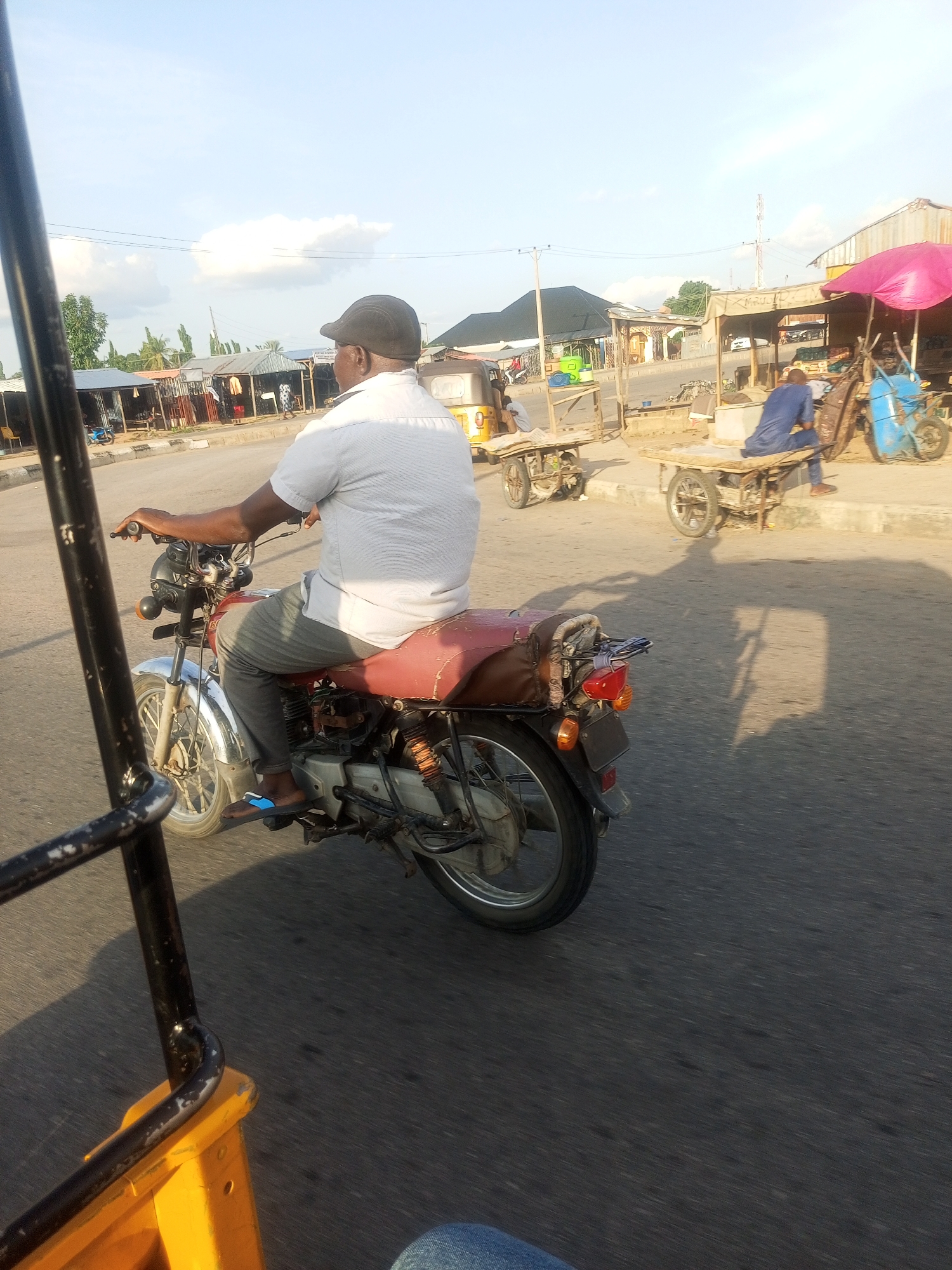 A man wearing a white short-sleeved shirt and gray pants rides a red and black motorcycle. He is also wearing a dark flat cap and blue and white sandals. The motorcycle is seen from the side, and the man is facing away from the camera. The scene appears to be a street in Jalingo, Nigeria, with shops and market stalls lining the road. In the background, there are several other people, some sitting on carts and others standing. A pink umbrella is visible over one of the stalls. The sky is bright, suggesting it is daytime, likely late afternoon due to the long shadows cast on the road. The weather appears to be clear and sunny. The image is captured from another vehicle, possibly a rickshaw or a similar mode of transport, as evidenced by the black metal frame in the foreground.