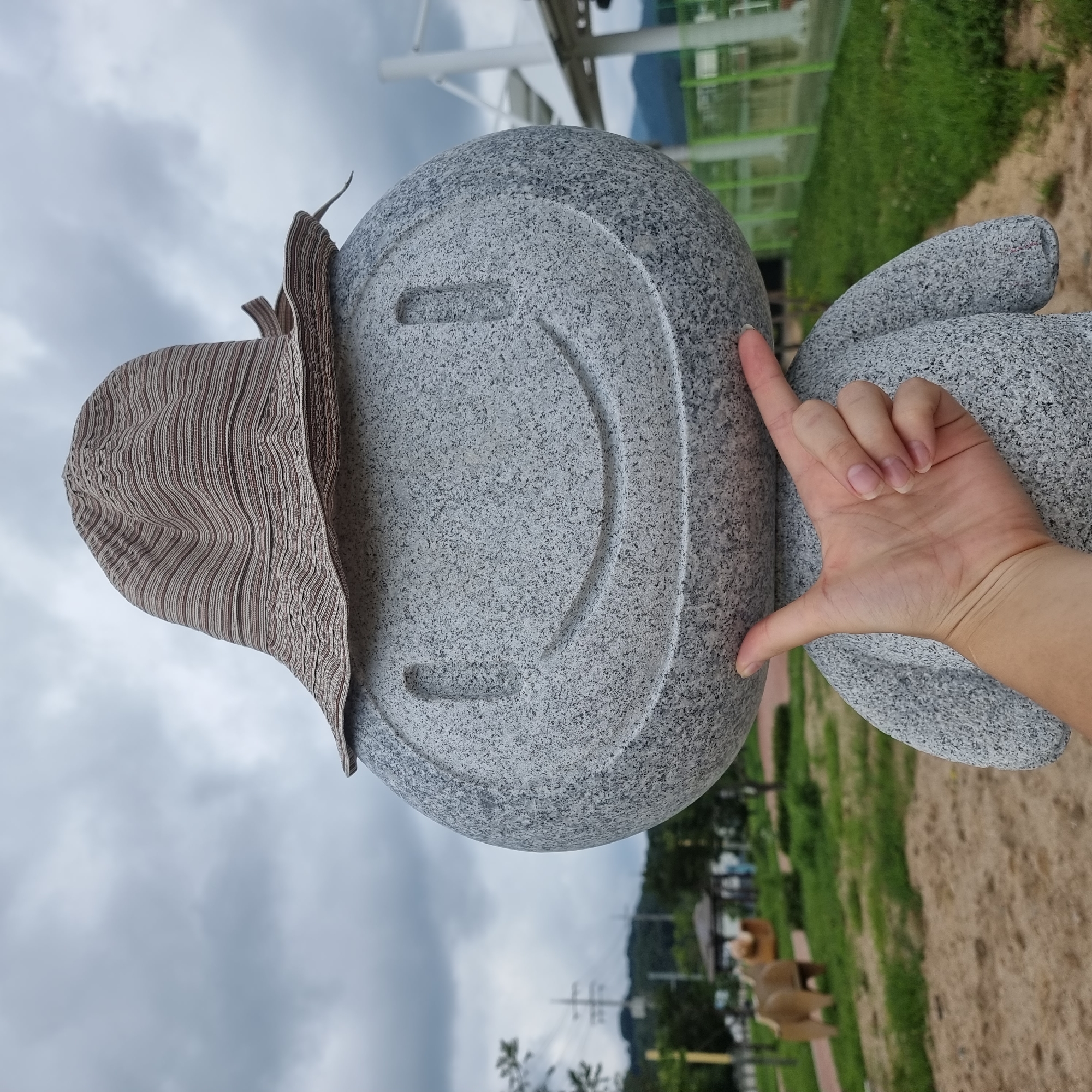 A gray stone statue resembling a round-headed figure with a carved smiley face and a brown and white striped hat is positioned outdoors. A person's left hand, with fingers extended, is touching the chin area of the statue, with the thumb and forefinger creating a gesture. The statue stands in a park-like setting with green grass, trees, a sandy path, and a blurred background that includes a small, wooden structure, power lines, and distant hills. The sky is overcast with gray clouds. The location is the Dunnae-myeon Sculpture Park in South Korea.