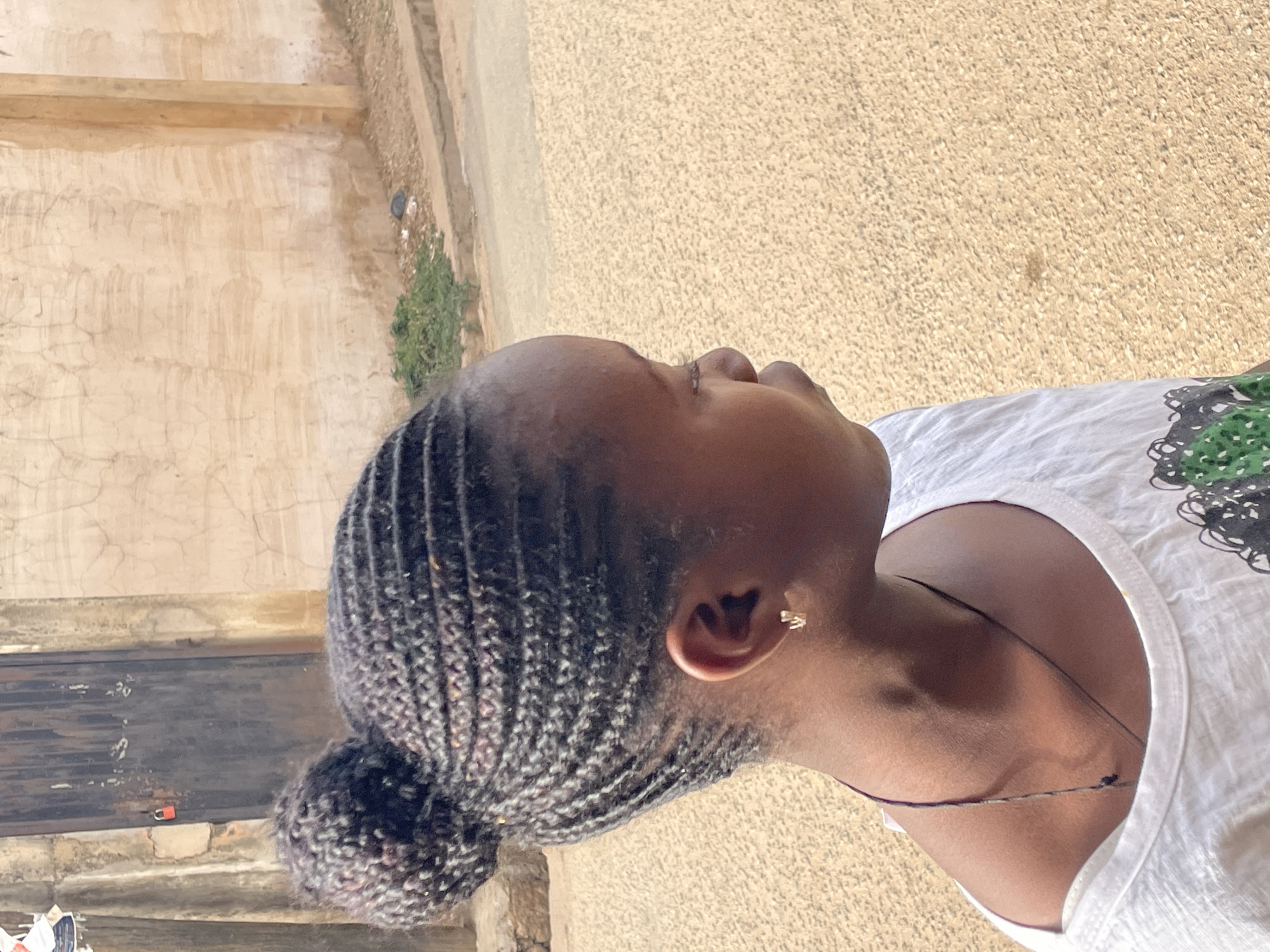 A close-up shot of a Black woman with braided hair pulled back into a bun. She is wearing a white t-shirt and a delicate necklace. Her head is tilted back, and her eyes are closed.
The background shows elements of an urban environment. A wall with a textured surface is on the left side of the frame. Behind the woman is what appears to be a paved area.
The image appears to be taken outdoors in Jos, Nigeria, based on the style of building and environmental cues.