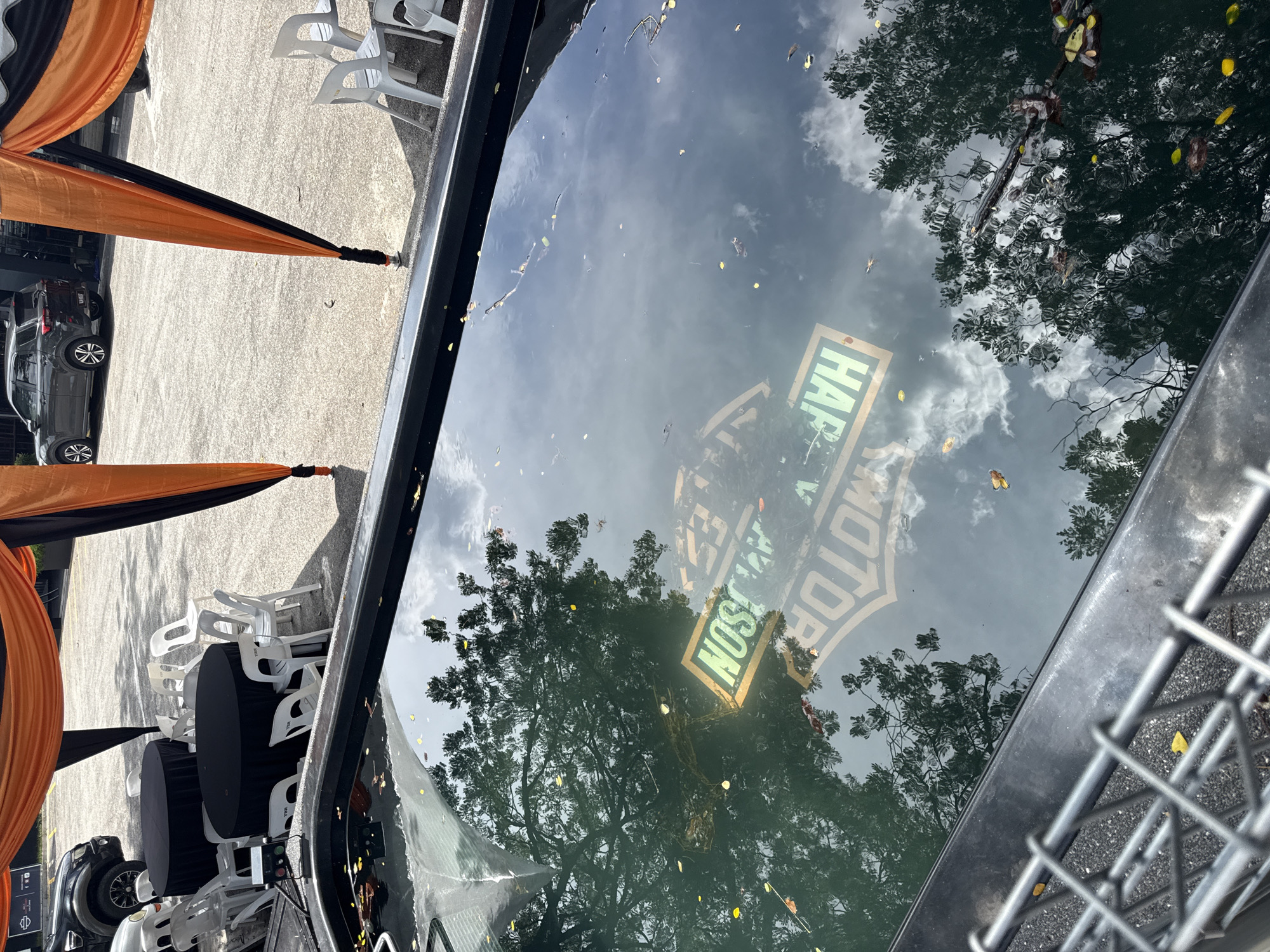 The image depicts a reflection of a Harley-Davidson sign and trees in what appears to be a car roof or another reflective surface. The scene is set outdoors, likely during the daytime, given the visible sky and tree foliage. The presence of black and orange draping suggests a possible event or themed setup. Tables and chairs are positioned to the side. The presence of cars and metal fencing add context to the scene, potentially indicating an urban or public space. Without additional data or reference points in the image, determining whether this is Kuala Lumpur, Malaysia, would be challenging.