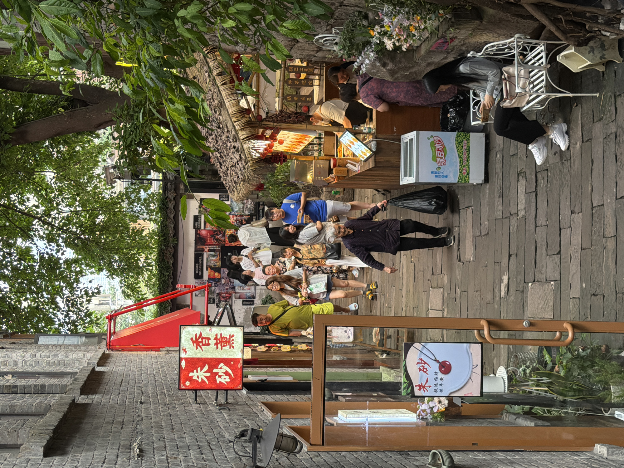 The image depicts a narrow, cobblestone alleyway in the Yuzhong District of China, likely during daytime given the ambient light. The alley is flanked by traditional brick buildings and features numerous shops and food stalls. In the foreground, a woman with long black hair is seated on an ornate white bench, holding a bowl and looking down at it. She is wearing a grey long-sleeved shirt and black pants, with white sneakers. To her right, a small white trash bin is visible. Adjacent to the bench is a white freezer with a colorful advertisement.
Further into the alley, a food stall with a thatched roof is open for business. A vendor, wearing a mask, is seen preparing food behind the counter. Several customers are gathered around the stall, some observing the offerings displayed on a digital screen. There are various food items and drinks being sold.
The alley is populated by a diverse group of people walking in both directions. Some are tourists, indicated by their attire and the presence of shopping bags. A man in a bright yellow t-shirt and green shorts is walking towards the foreground, while a group of women in casual wear are seen further down the alley. Red lanterns and traditional Chinese signage are visible on the buildings, adding to the cultural ambiance. Lush green foliage from trees overhangs parts of the alley, casting shadows and contributing to the atmosphere. The overall scene suggests a vibrant and bustling market or street.