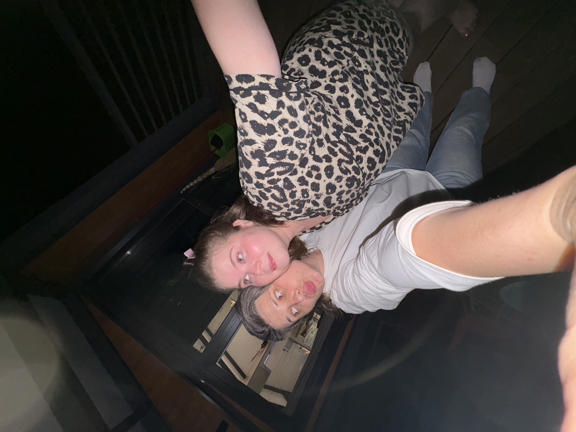 The media file shows two people, likely a mother and daughter, lying down and taking a selfie. The younger person, on top, is wearing a leopard-print top and has a pink bow in her hair. Her eyes are wide and looking upwards. The older person, below, is wearing a white t-shirt. Her hair is grey and pulled back, and she is sticking her tongue out, making a silly face. They are on a wooden deck or porch, indicated by the wooden planks and railing in the background. The scene appears to be outdoors at night, as suggested by the darkness and artificial lighting. The overall mood is playful and casual. The reflection on the surface below them suggests they might be on a glass table or floor.