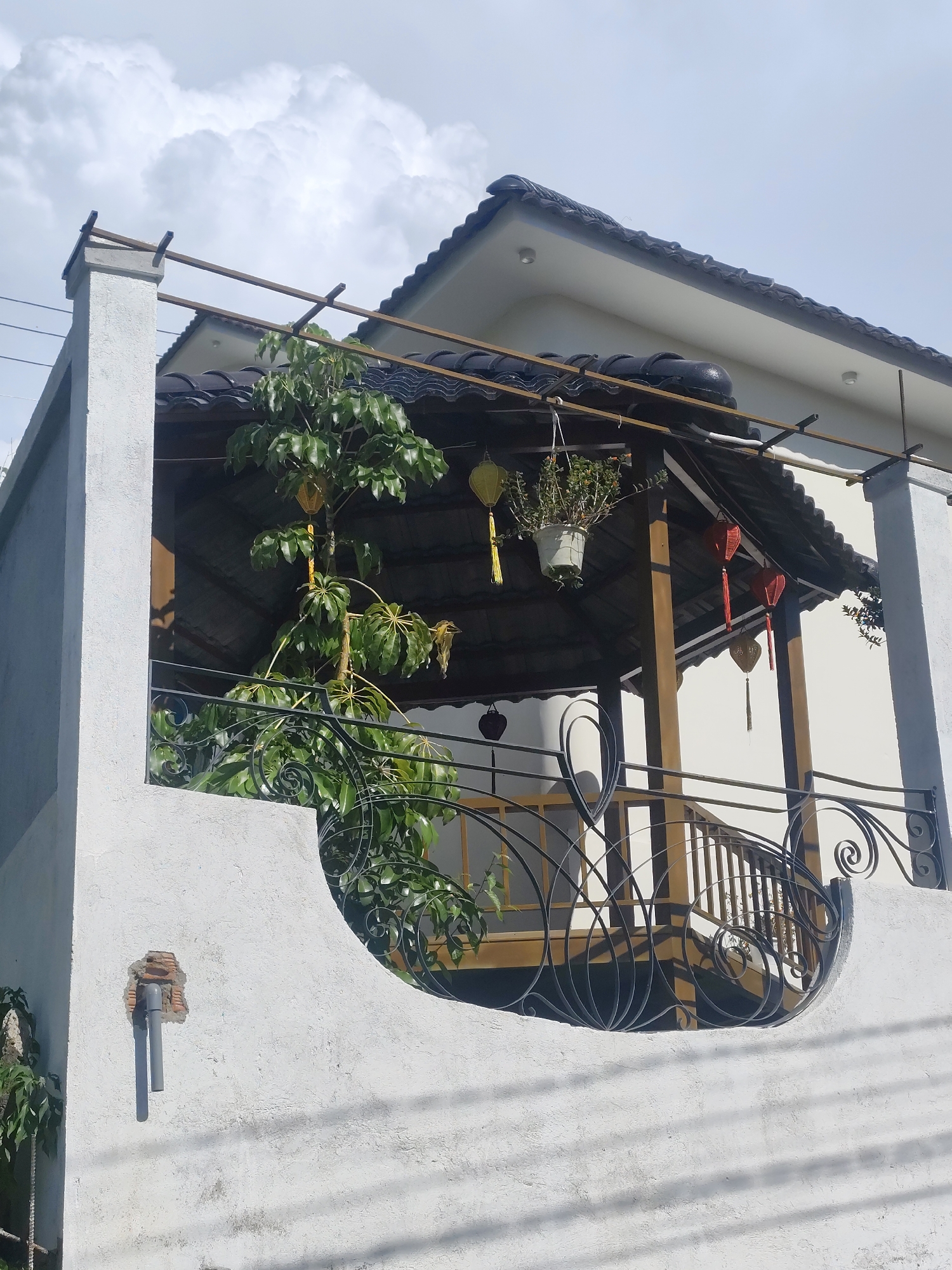 The image displays a close-up view of a balcony on a white building with a dark tiled roof, likely located in Lâm Viên - Đà Lạt, Vietnam. The balcony features ornate black metal railings with swirling designs. A large green plant with broad leaves is growing on the balcony, its branches reaching upwards. Above the balcony, under a covered roof, several colorful lanterns hang, including yellow and red ones. One yellow lantern is visible on the left, and two red lanterns are suspended on the right. A potted plant with green foliage is also seen hanging from the covered roof. The building's exterior wall has a rough, textured finish and shows signs of wear, including a section where bricks are exposed around two grey pipes protruding from the wall. The sky above is partly cloudy with large, white cumulus clouds against a pale blue sky. The scene is captured from a low angle, looking up at the balcony and the building.