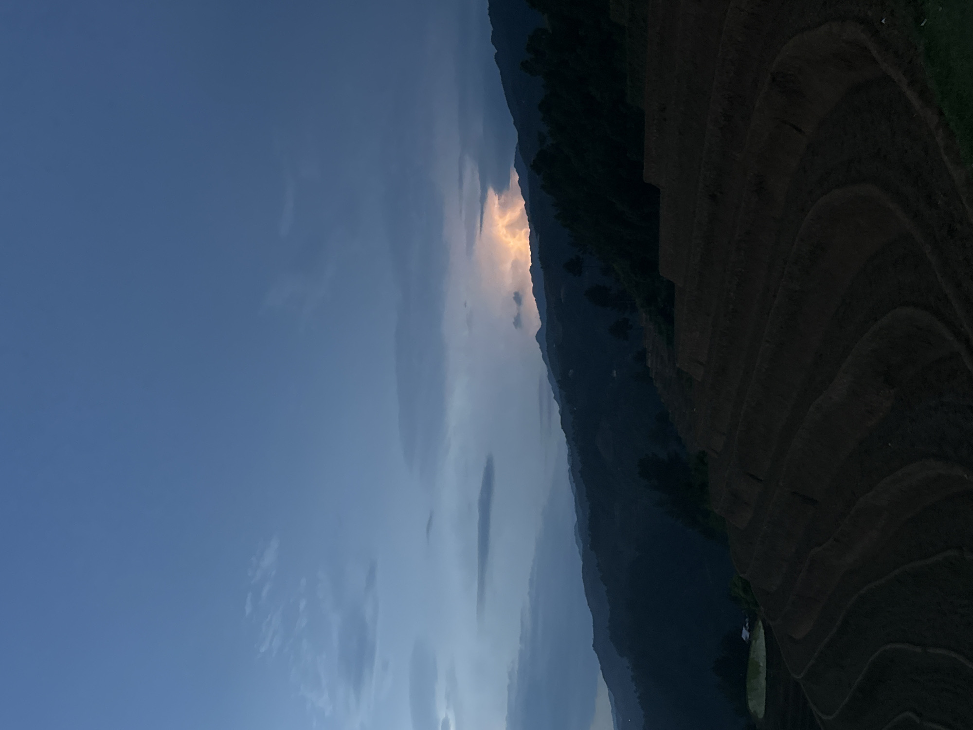 The image captures a majestic mountainous landscape at twilight, likely during dusk or dawn. The scene is dominated by a vast sky, which transitions from a deep blue at the top and left to a lighter hue towards the center. Scattered clouds are visible, with a striking, warm glow of orange and yellow emanating from behind a mountain ridge, illuminating a section of the sky and suggesting the sun is low on the horizon.
Across the middle ground, towering mountains rise in dark silhouettes, their rugged contours sharply defined against the dramatic sky. To the right, expansive terraced fields are visible, showcasing their distinct, curving patterns as they descend the mountain slopes. These terraces are mostly in shadow, emphasizing the low light conditions. In the lower right corner, a small, lighter-colored area suggests a body of water or a flooded section within the terraced landscape.
No people or explicit activities are visible, presenting a tranquil and awe-inspiring natural vista. The overall mood is one of serene grandeur and natural beauty, characteristic of mountainous rice paddy regions such as Mù Cang Chải, Vietnam. There is no visible text.
