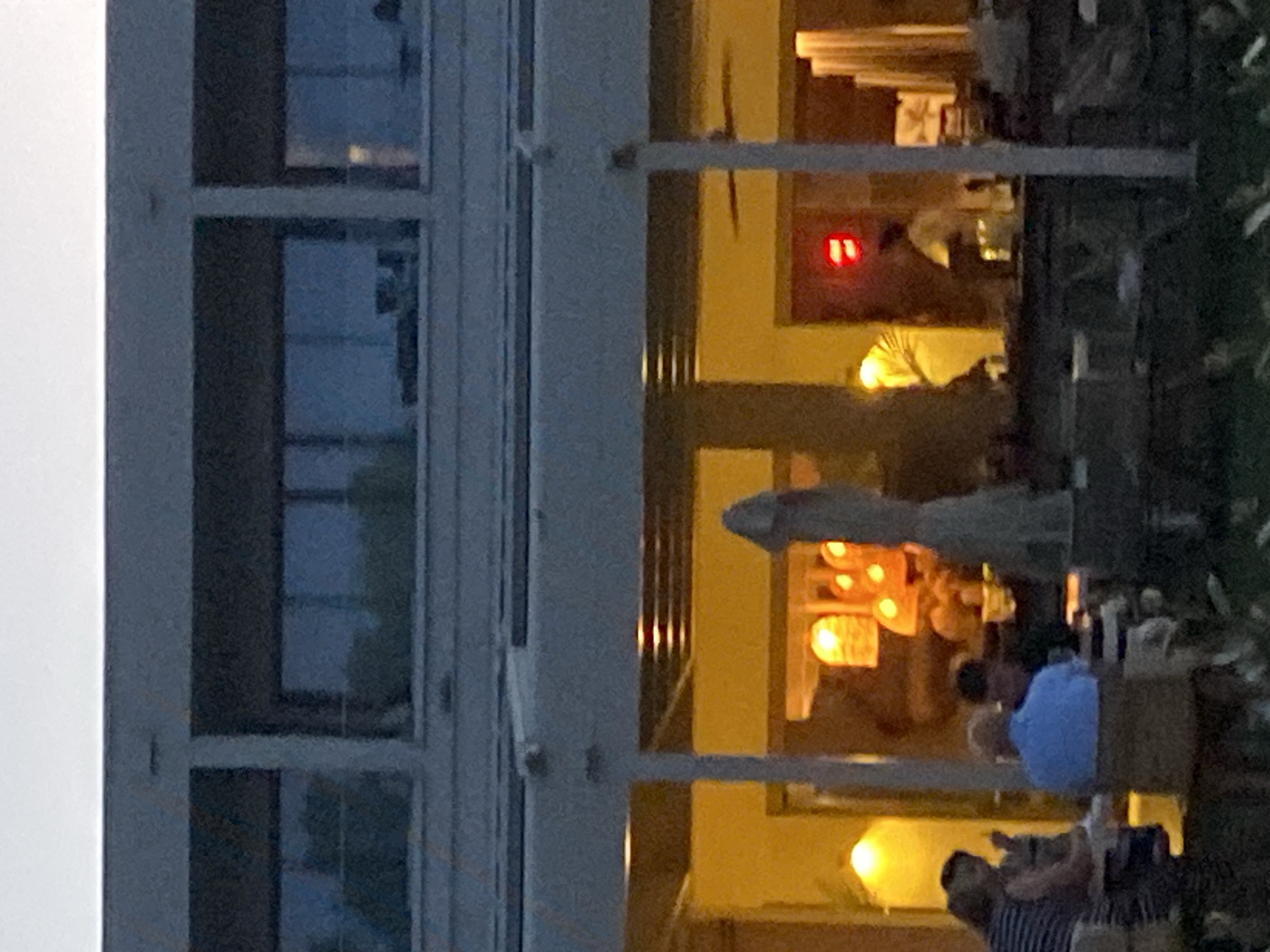 The image shows a restaurant with outdoor seating. Several people are dining at tables under a covered patio. The restaurant is lit with warm yellow lights, and there are also pendant lights hanging from the ceiling. In the background, there is a red digital clock displaying "11". The architecture of the building appears modern, with large windows and a clean, minimalist design. The overall atmosphere seems relaxed and inviting, suggesting a pleasant dining experience. Without further information, it is impossible to pinpoint the exact location within Estepona, Spain.