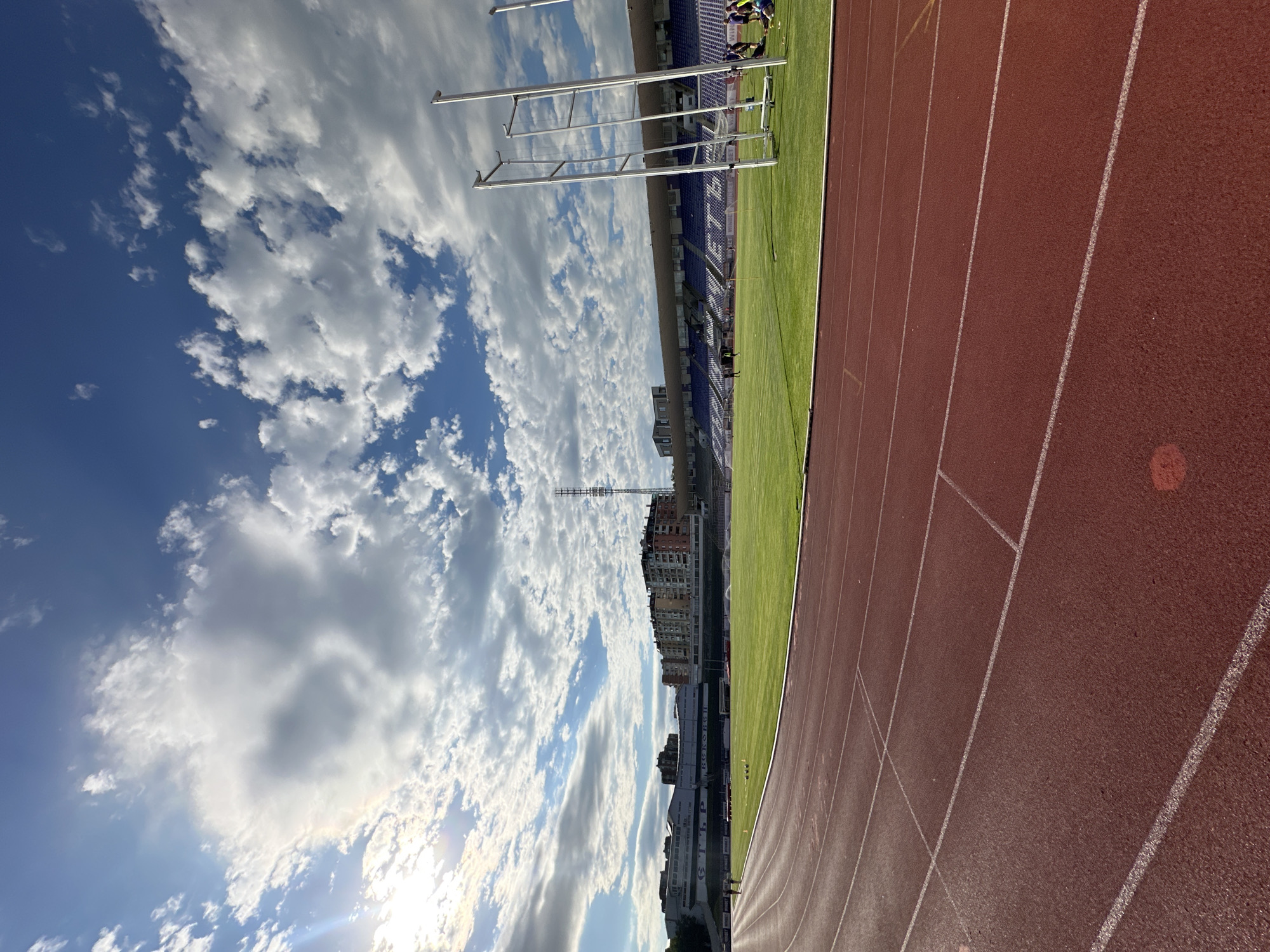 This is a vertical, eye-level, medium-wide shot of the Ivaylo Stadium track and field in Veliko Tarnovo, Bulgaria. The stadium's red track with white lane markings takes up the lower half of the frame, curving from left to right. Beyond the track is a vibrant green grass field with scattered white lines. In the middle ground, a row of empty blue stadium seats is visible, topped by a white roof. Behind the seats, several multi-story residential buildings and a telecommunications tower are visible against a bright blue sky filled with fluffy white clouds. The sun shines from the upper left, casting a warm light on the scene. Two sets of tall, silver upright poles with horizontal bars, likely for high jump or pole vault, are positioned on the right side of the field. Several people, appearing as small dark figures, are scattered across the field and near the stadium seats, engaged in various activities.