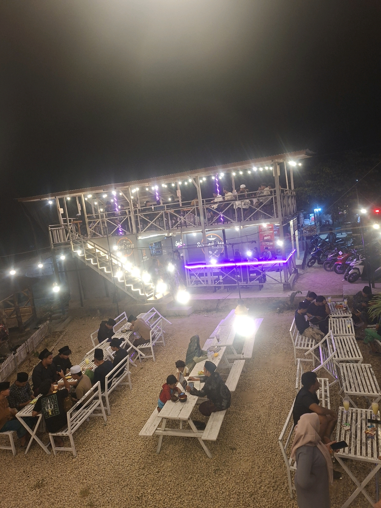 The image captures an outdoor seating area at night, likely a restaurant or cafe, located in Lantek Barat, Indonesia. The scene is illuminated by string lights and spotlights, casting a warm glow on the gravel-covered ground and the predominantly wooden structures.
In the foreground, several white wooden picnic tables with benches are occupied by groups of people. Some individuals are seated, engaged in conversation, using their phones, or looking at laptops. To the left, a group of men wearing peci (traditional caps) are gathered around a table, some eating or drinking. Further to the right, a child is seated at a picnic table, seemingly focused on a bowl.
A multi-level wooden building with a slanted roof is visible in the mid-ground. The upper level has a balcony with several patrons seated. A prominent staircase with illuminated steps leads up to this level. The lower level of the building features a stage area with purple lighting and a sign that appears to read "Super Jorit" with other text underneath.
To the right of the building, a row of parked motorcycles, mostly scooters, is visible. Some of these are red and white. In the background, trees and foliage can be seen, suggesting a somewhat rural or suburban setting. The overall atmosphere appears casual and social, with people enjoying an evening out.