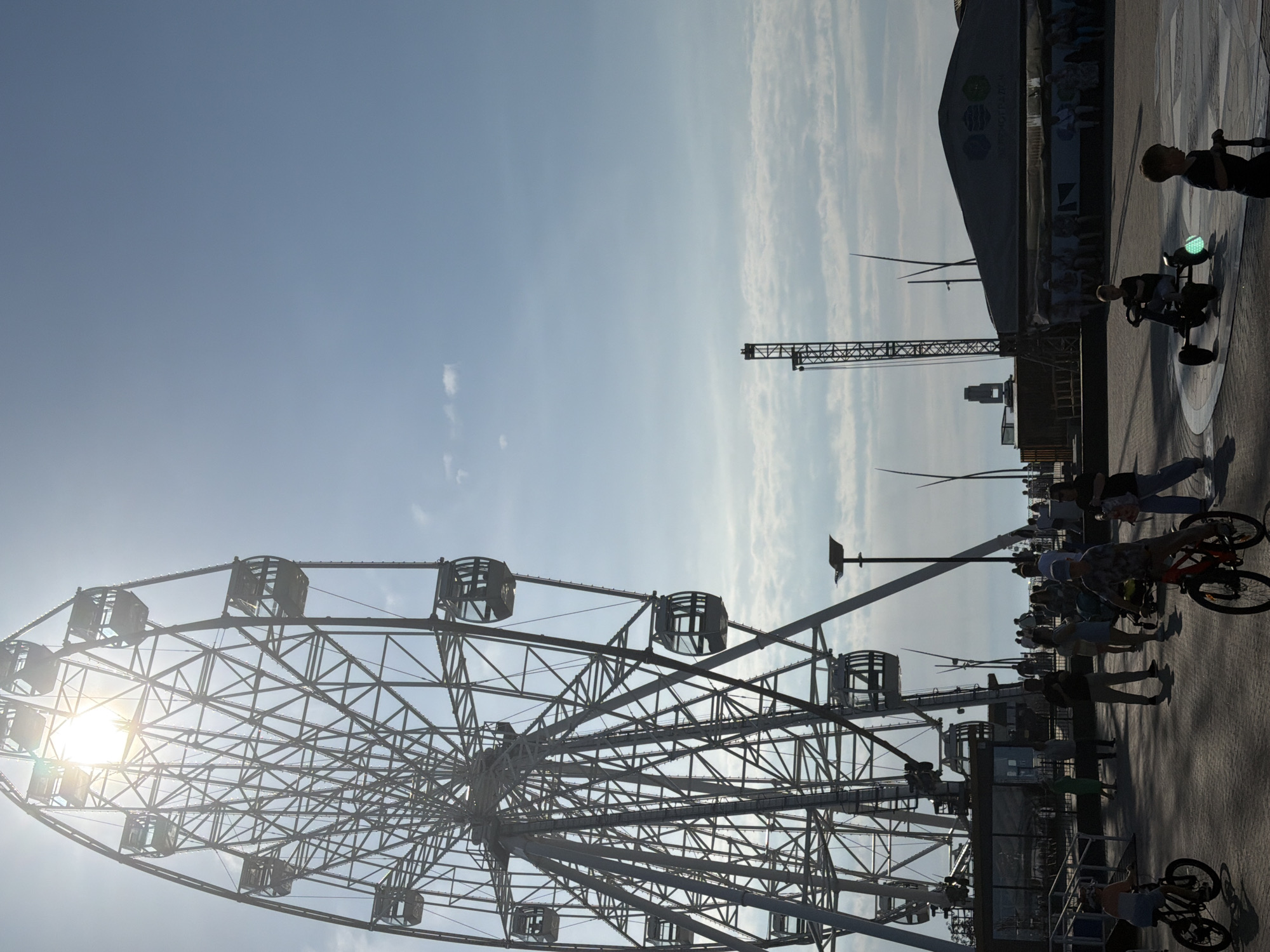 In Zelenogradsk, Russia, the image captures a bustling scene in an amusement park or waterfront area on a bright, sunny day. A towering Ferris wheel dominates the left side of the frame, its intricate metal structure silhouetted against a clear blue sky, with the sun's rays beaming through its spokes. Below the Ferris wheel, a paved area is populated by people enjoying various activities. In the foreground, two children are on bicycles, one in the lower left corner and another in the lower center, pedaling across the pavement. To the right of the children on bicycles, two boys are engaged with wheeled toys; one is seated in a pedal-powered vehicle with a green light, and the other stands beside him, holding onto a scooter-like device. Scattered throughout the scene are other individuals, some standing and conversing, others strolling, and a group is seated under a large, dark canopy structure in the mid-ground. A tall, lattice-like structure, possibly a construction crane or another amusement ride, stands to the right of the Ferris wheel, further emphasizing the recreational nature of the location. The overall impression is one of a lively public space filled with families and visitors taking advantage of pleasant weather.