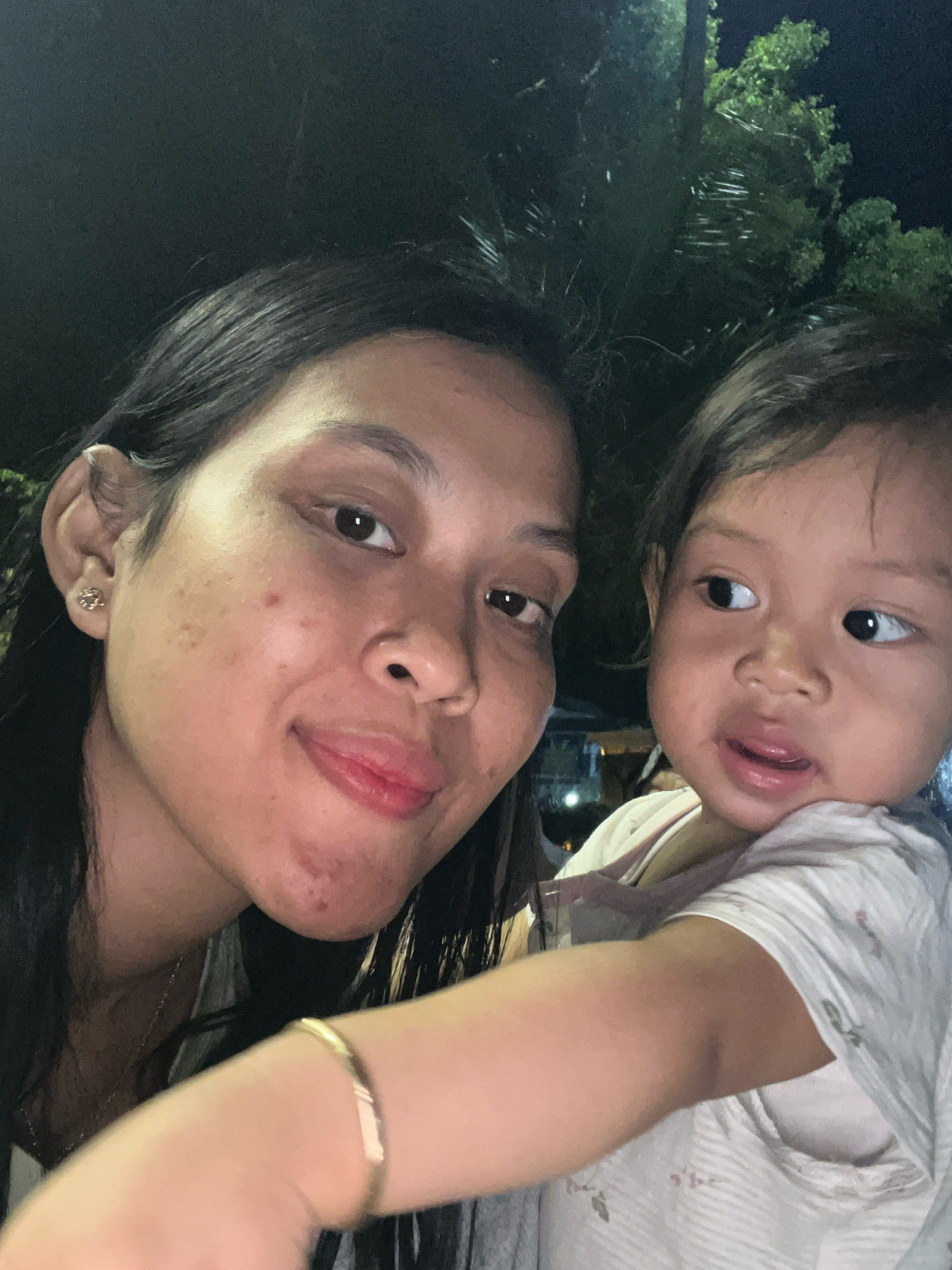 A close-up selfie, taken at night, features two individuals in the foreground. On the left is an adult woman with long, dark hair, looking directly at the camera with a slight smile. She is wearing an earring in her left ear. To her right, and slightly behind her, is a young child with dark hair, looking to the right. The child is wearing a light-colored, patterned shirt. The background is dark, with some indistinct foliage visible, suggesting an outdoor setting at night, potentially in Kecamatan Pati, Indonesia, as indicated by the provided context.
