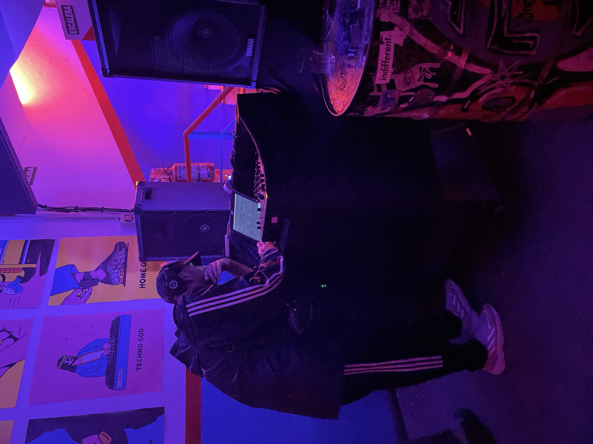 A medium, rotated shot shows a DJ, dressed in an Adidas tracksuit, performing at a venue in Buenos Aires, Argentina. The DJ wears a cap and headphones and is seen behind a DJ booth, which is draped in black cloth. Above the booth are two Yamaha speakers. To the right of the DJ booth is a metal barrel. The venue is lit with purple and blue lighting. On the wall to the left of the DJ, there are posters of various designs. The shot also includes signs labeled "ESCALERA" and "SALIDA."