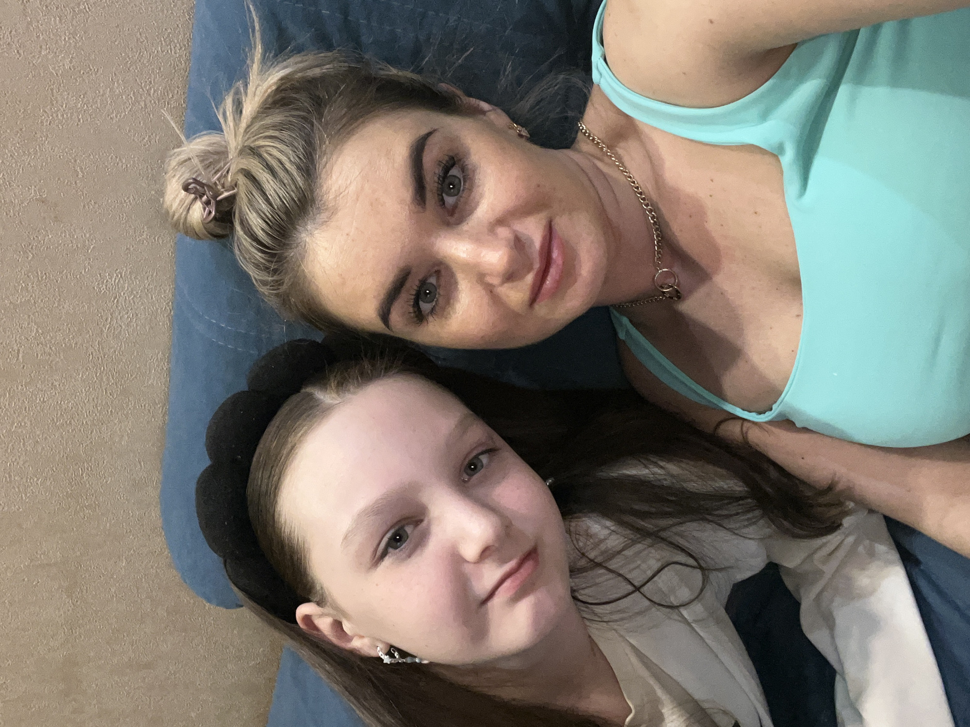 Two light-skinned females are lying together on a blue surface, likely a couch or bed. The adult woman is positioned at the top, wearing a light blue sleeveless top and a gold chain necklace. Her blonde hair is styled in a bun. The younger girl, presumably a child, is below her, wearing a black headband and a white garment. Both are facing upwards, looking at the camera. The location cannot be determined from the image.