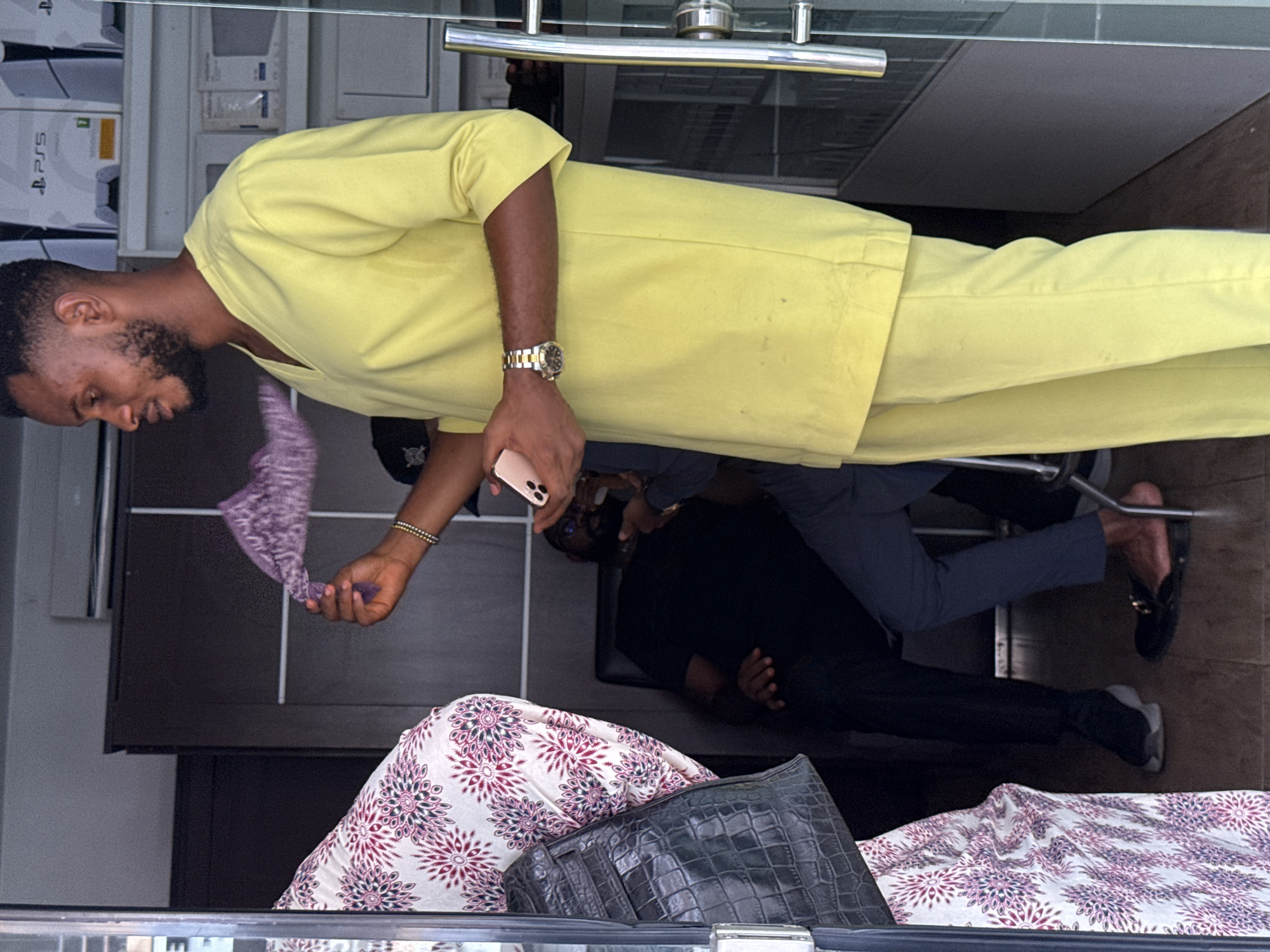 A man in a yellow suit is standing indoors, potentially in a store based on the reflected display cases containing Playstation 5 consoles visible in the top left. He holds a pink and purple patterned cloth in one hand and a smartphone in the other. He is wearing a gold watch and beaded bracelet.
Several other people are present in the scene. One man sits on a chair. Another man is on the floor taking a photo of a handbag sitting on a printed fabric.
The photo was taken in Abuja, Nigeria.