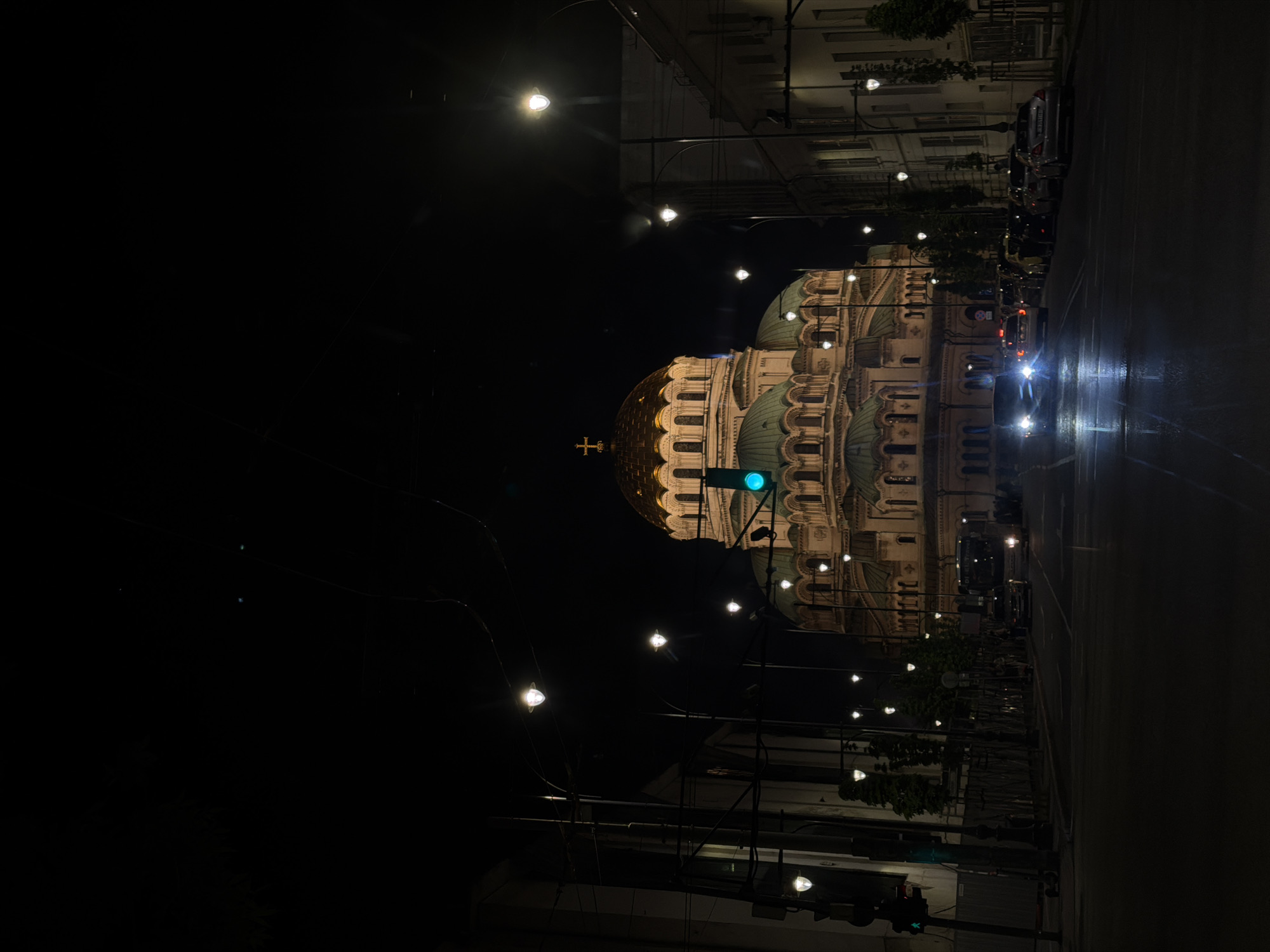 This nighttime shot depicts the Alexander Nevsky Cathedral in Sofia, Bulgaria, prominently illuminated. The street on the right side of the frame shows cars and streetlights reflecting off the wet pavement. The cathedral's domes are a mix of gold and green hues, and a gold cross crowns the central dome. A green traffic light is also visible. The left side of the image is dark, with some streetlights and wires silhouetted against the night sky. The photograph is taken at street level, with the camera oriented vertically.