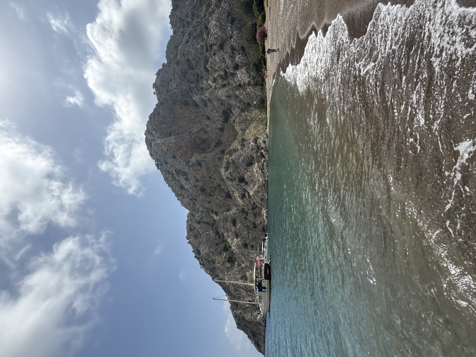 The image shows a scene at a beach in Kumluca, Turkey, with a large, rocky mountain rising sharply along the shoreline. The beach is narrow, with dark sand. The water is clear and turquoise, with waves gently lapping onto the shore. A few boats are docked against the base of the mountain. A single person is standing on the beach near some flowering plants. The sky is light blue with scattered white clouds.