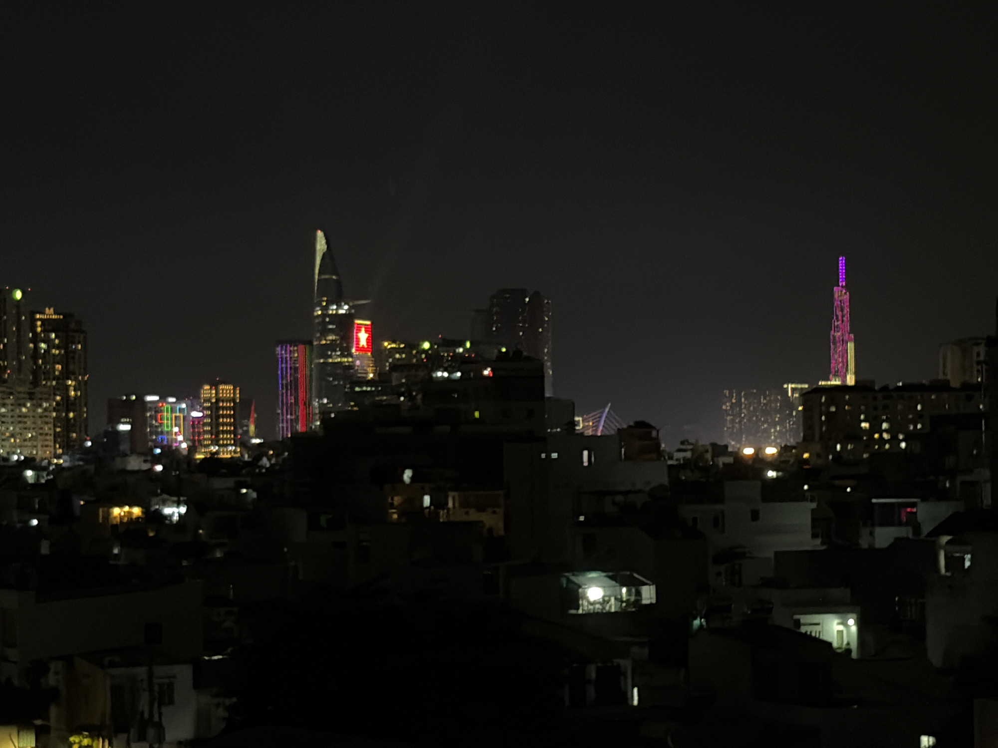 This is a nighttime aerial photograph of a city skyline in Thủ Đức, Vietnam. The foreground is dominated by the dark silhouettes of buildings, with some illuminated windows scattered throughout. In the mid-ground, more brightly lit buildings are visible, some with colorful architectural lighting. Several distinct skyscrapers stand out in the background, including one with a prominent red star on its facade and another with a tall, slender, illuminated spire. The overall impression is of a bustling urban environment at night, with a dense concentration of high-rise buildings. The sky is dark, suggesting it is late at night or early morning.